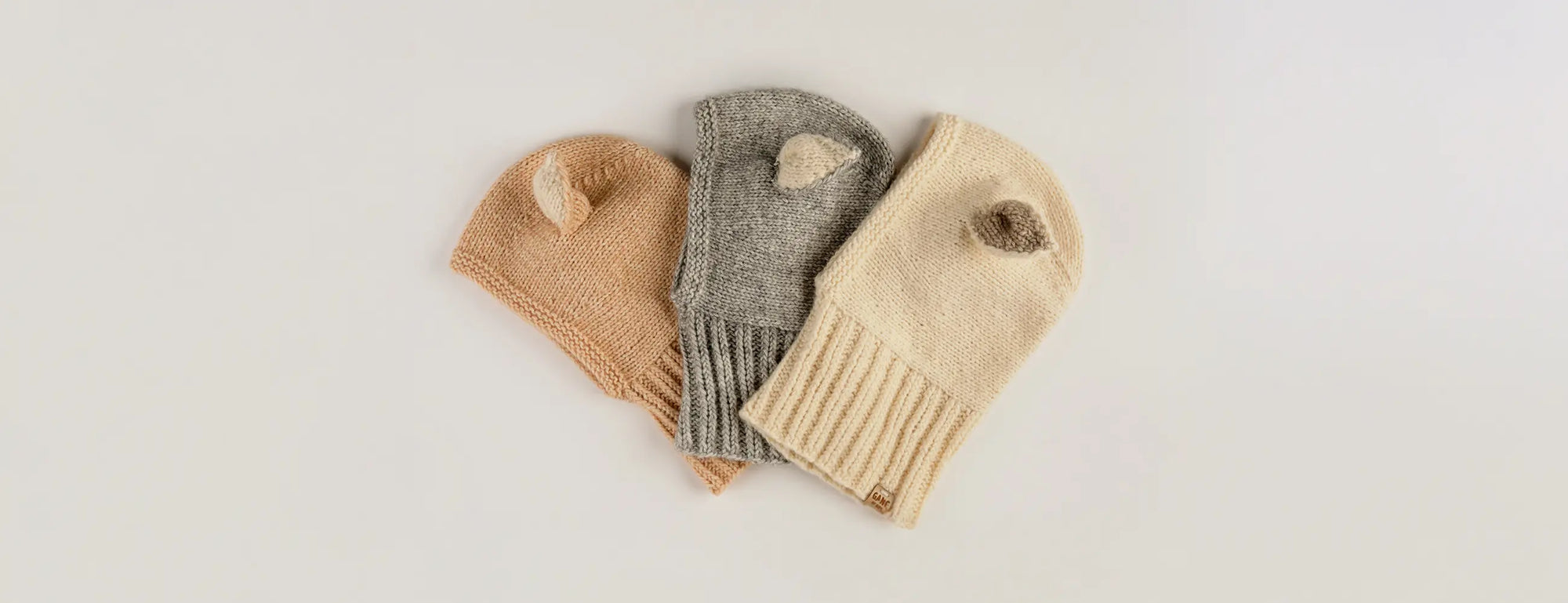 Three Sheep Hat cotton bonnets with ears, in peach, grey and cream, laid flat side by side.