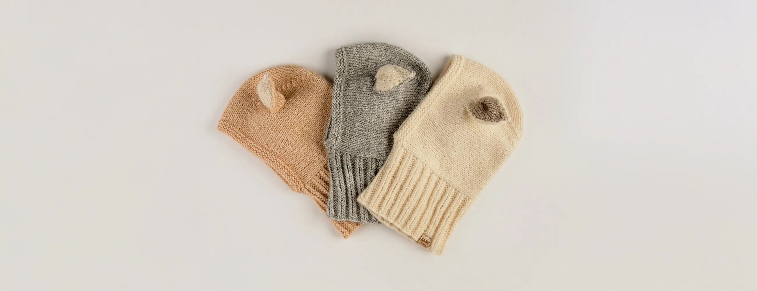Three Sheep Hat cotton bonnets with ears, in peach, grey and cream, laid flat side by side.