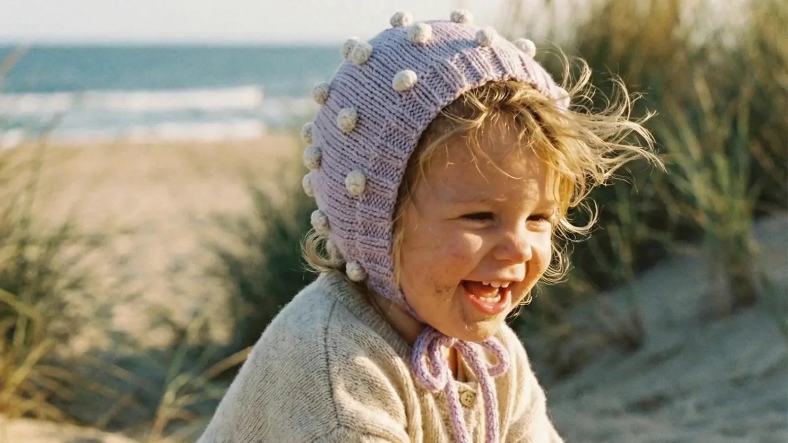 Smiling baby on a sandy beach wearing a hand-knitted lavender bonnet with popcorn texture