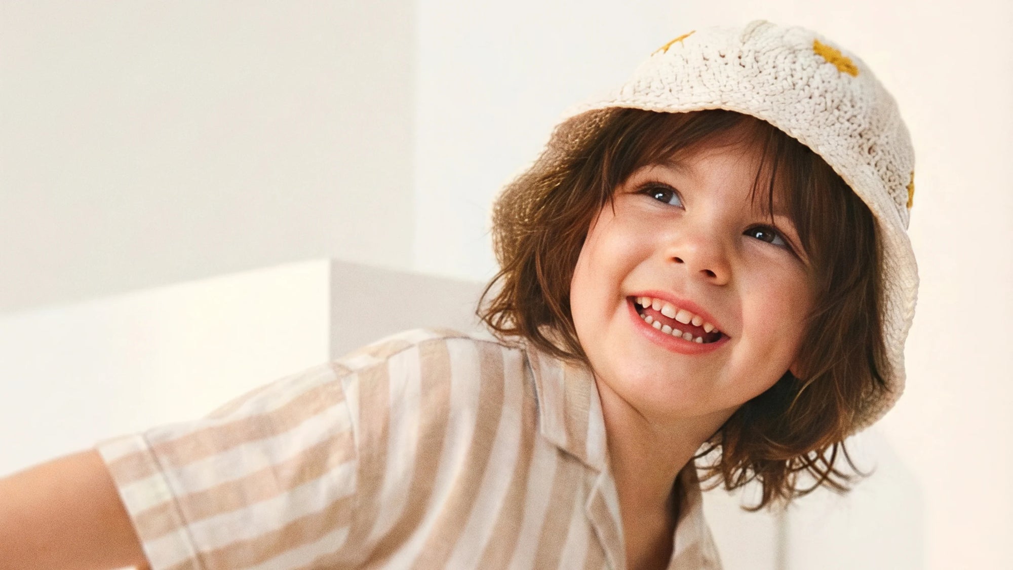 Toddler girl wearing an amber daisy sun hat with white flower pattern, sitting by a sunny window