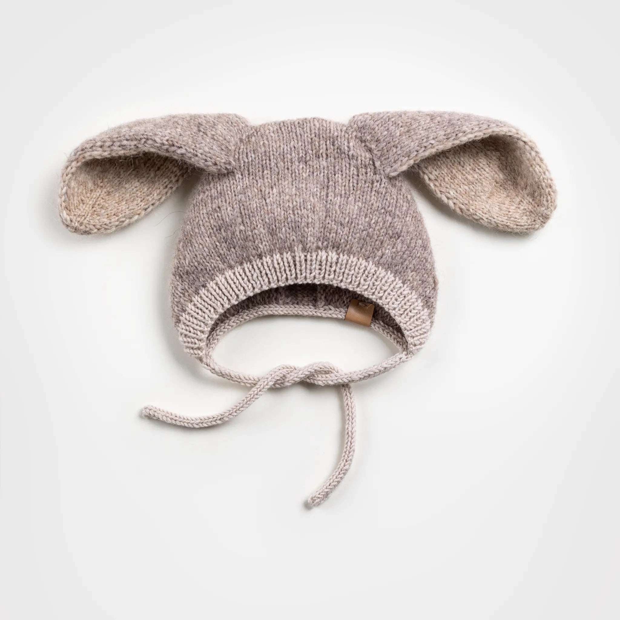 Flat lay of a handmade light brown and beige knitted baby bonnet with floppy bunny ears and chin ties, placed against a solid white background.