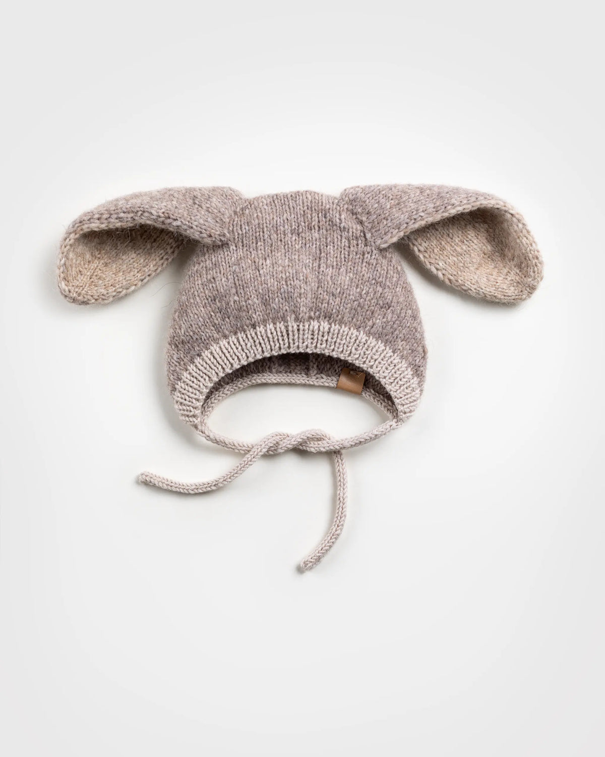 Flat lay of a handmade light brown and beige knitted baby bonnet with floppy bunny ears and chin ties, placed against a solid white background.