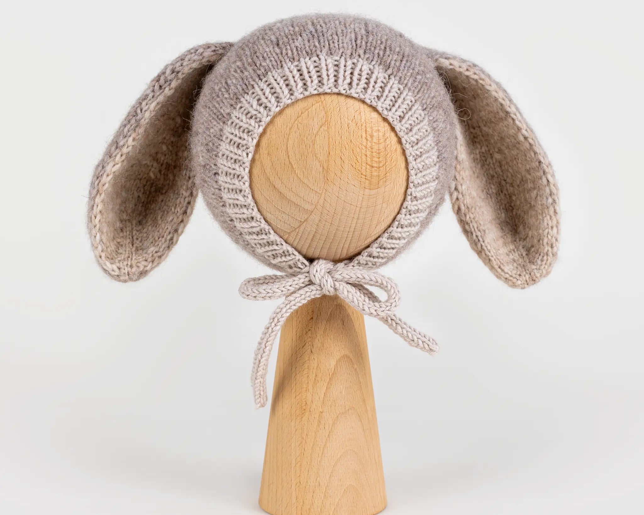Front view of a handmade light brown knitted baby bonnet with long bunny ears and chin ties, displayed on a wooden mannequin head against a white background.