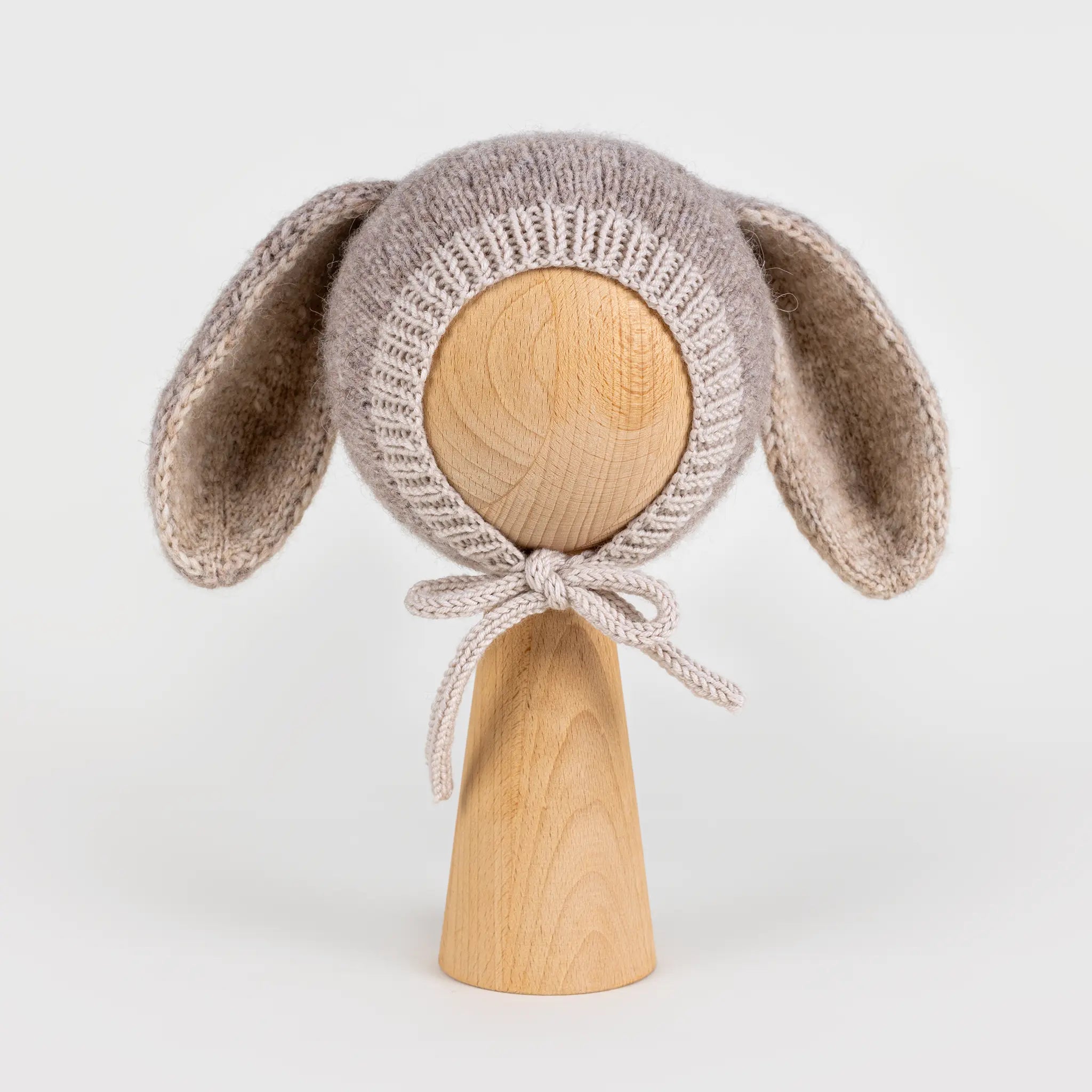 Front view of a handmade light brown knitted baby bonnet with long bunny ears and chin ties, displayed on a wooden mannequin head against a white background.