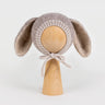 Front view of a handmade light brown knitted baby bonnet with long bunny ears and chin ties, displayed on a wooden mannequin head against a white background.