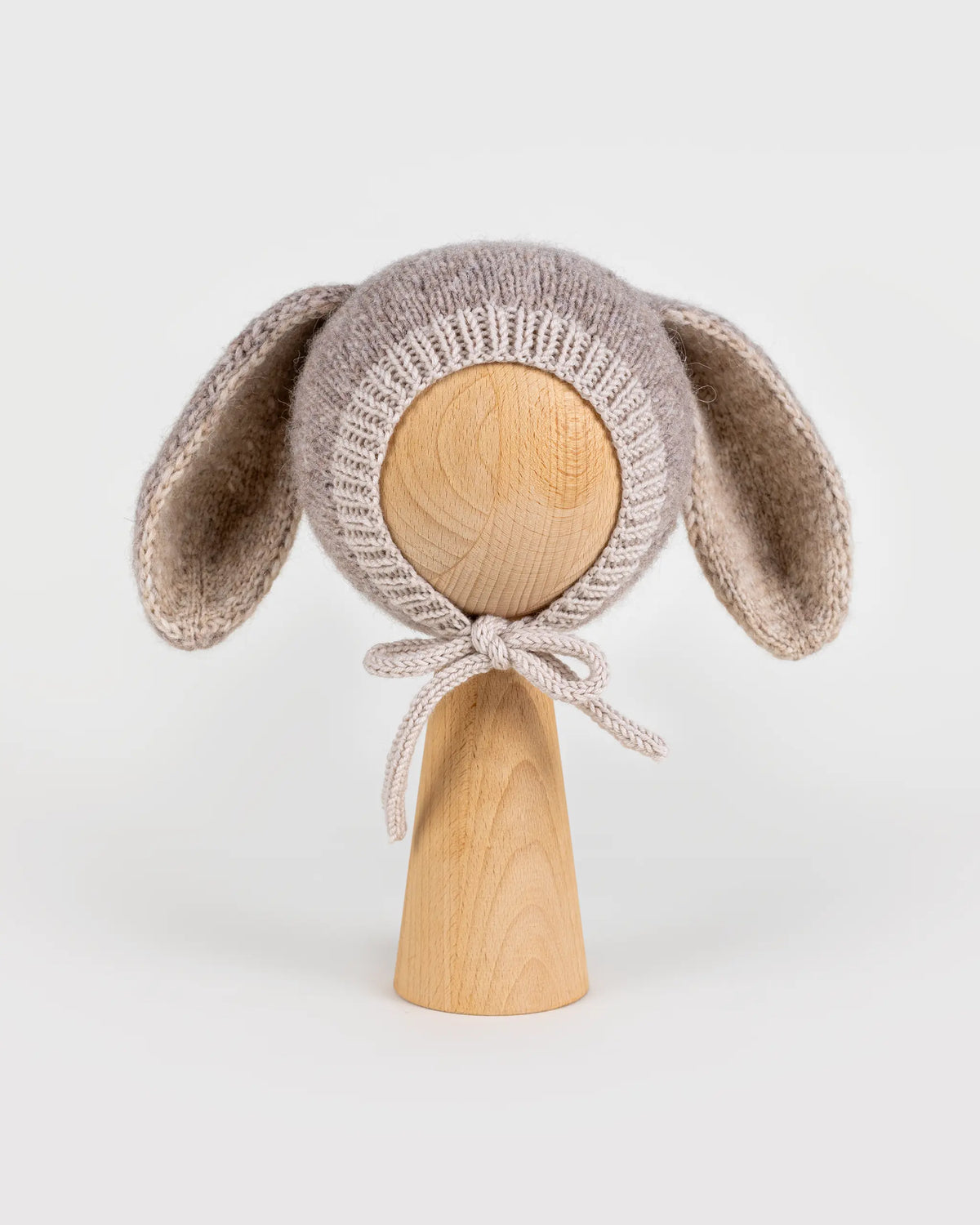 Front view of a handmade light brown knitted baby bonnet with long bunny ears and chin ties, displayed on a wooden mannequin head against a white background.