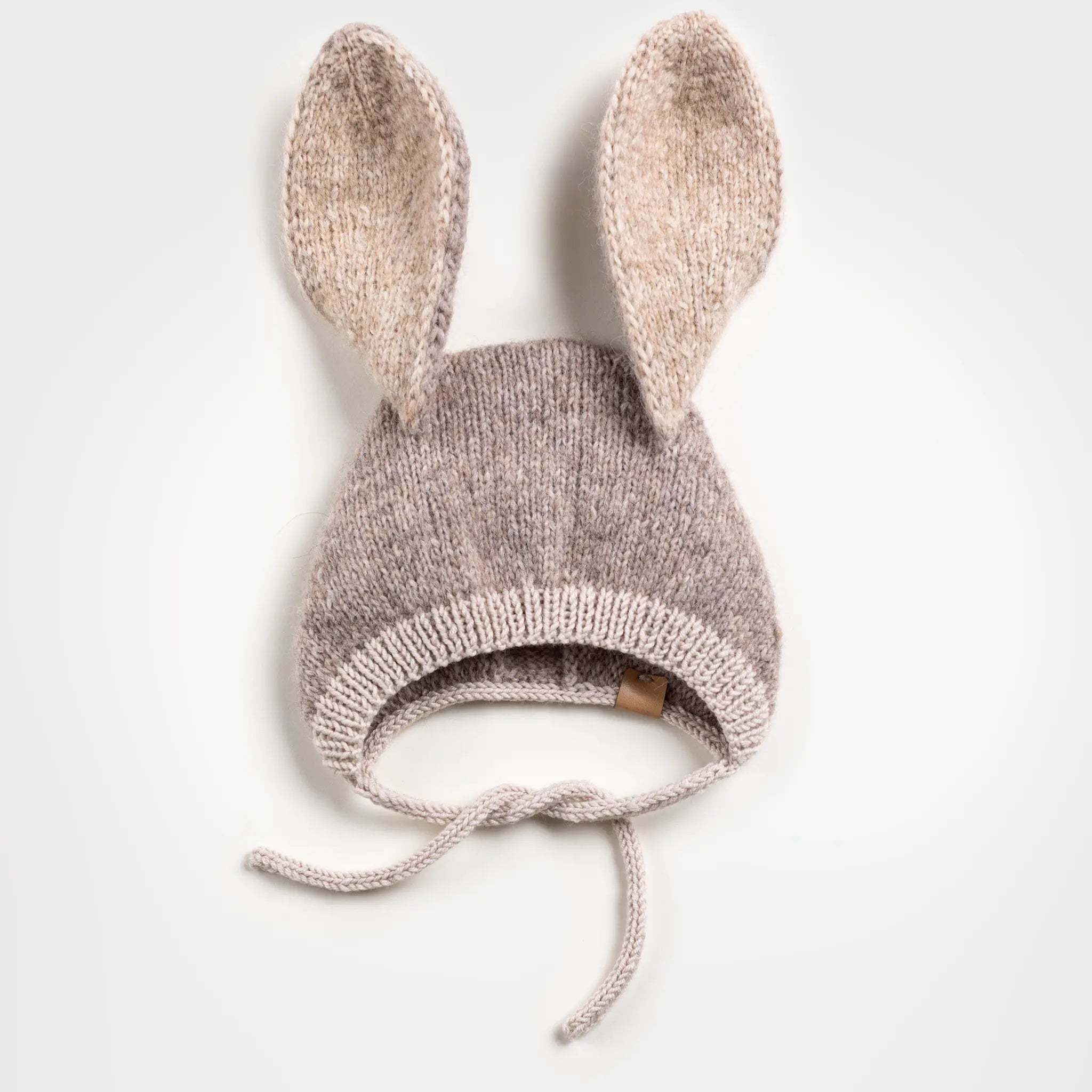 Flat lay of a handmade brown and beige knitted baby bonnet with long, upright bunny ears and chin ties, positioned against a plain white background.