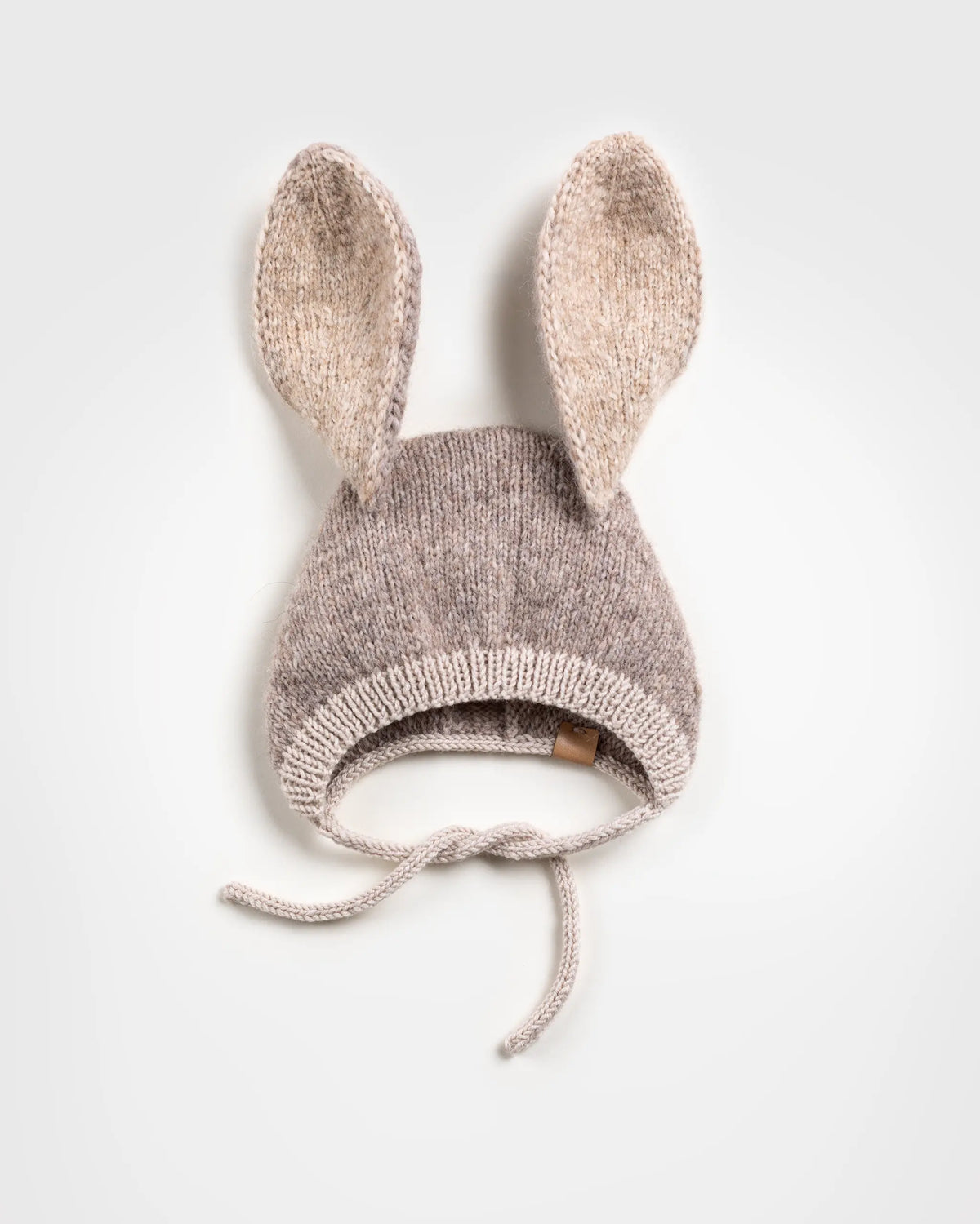 Flat lay of a handmade brown and beige knitted baby bonnet with long, upright bunny ears and chin ties, positioned against a plain white background.