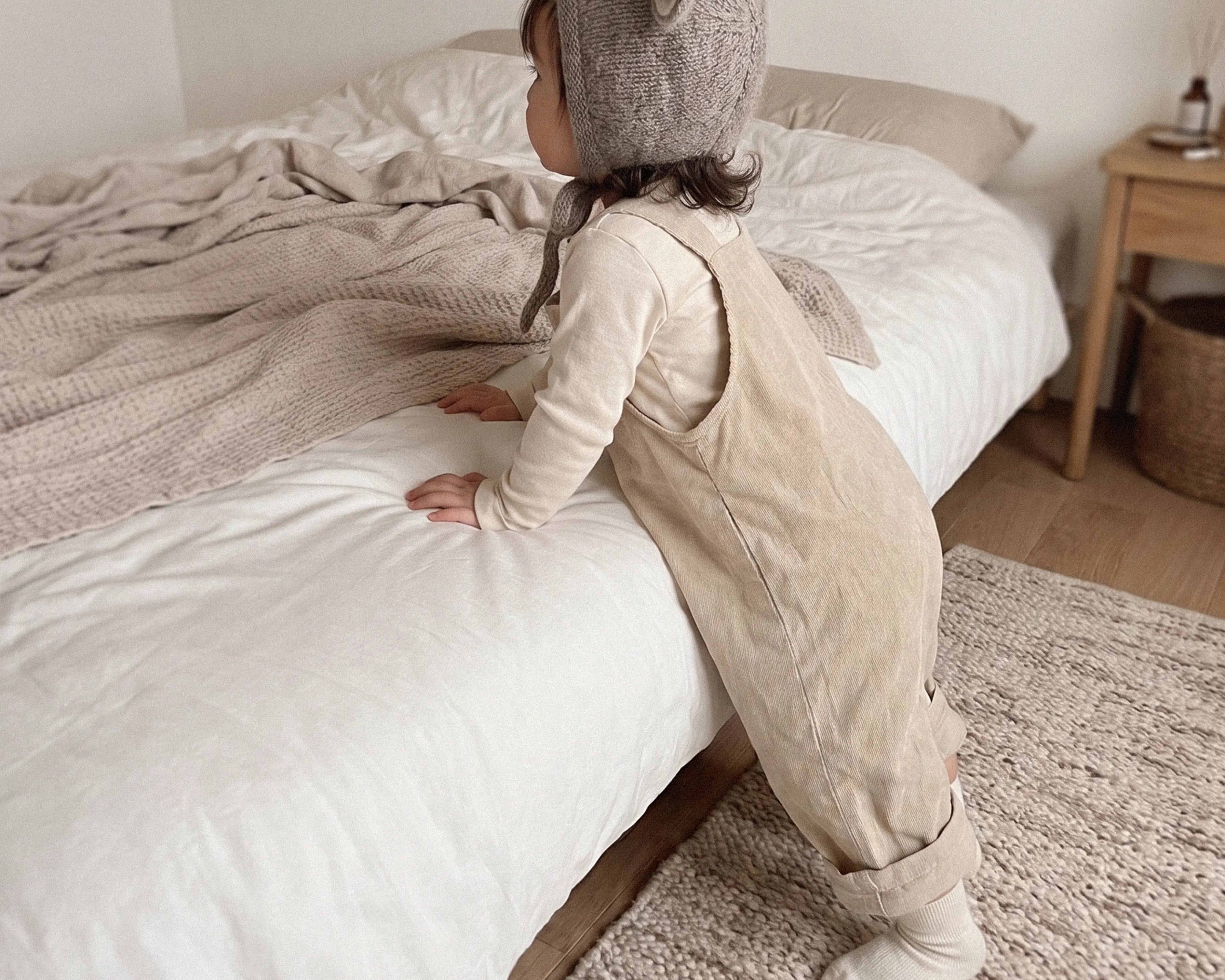 A toddler stands on a textured rug beside a bed, wearing a beige corduroy overall-style romper with a gray knit animal hood featuring white ears. The room has neutral beige and white bedding, a wooden side table, and a light rug.