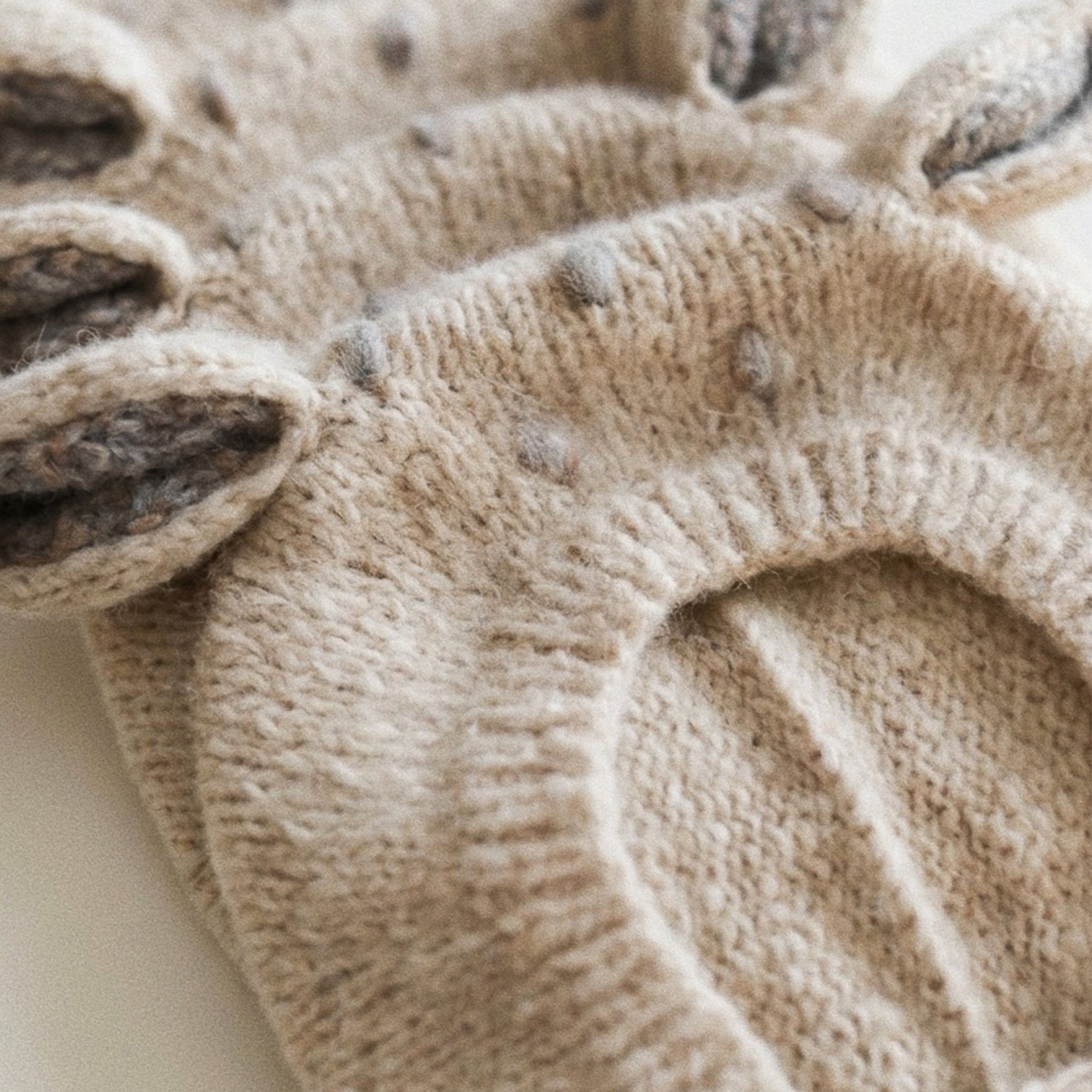 Close-up of a beige knit pet hood with ear flaps and small gray pom-pom accents, photographed on a light neutral background.