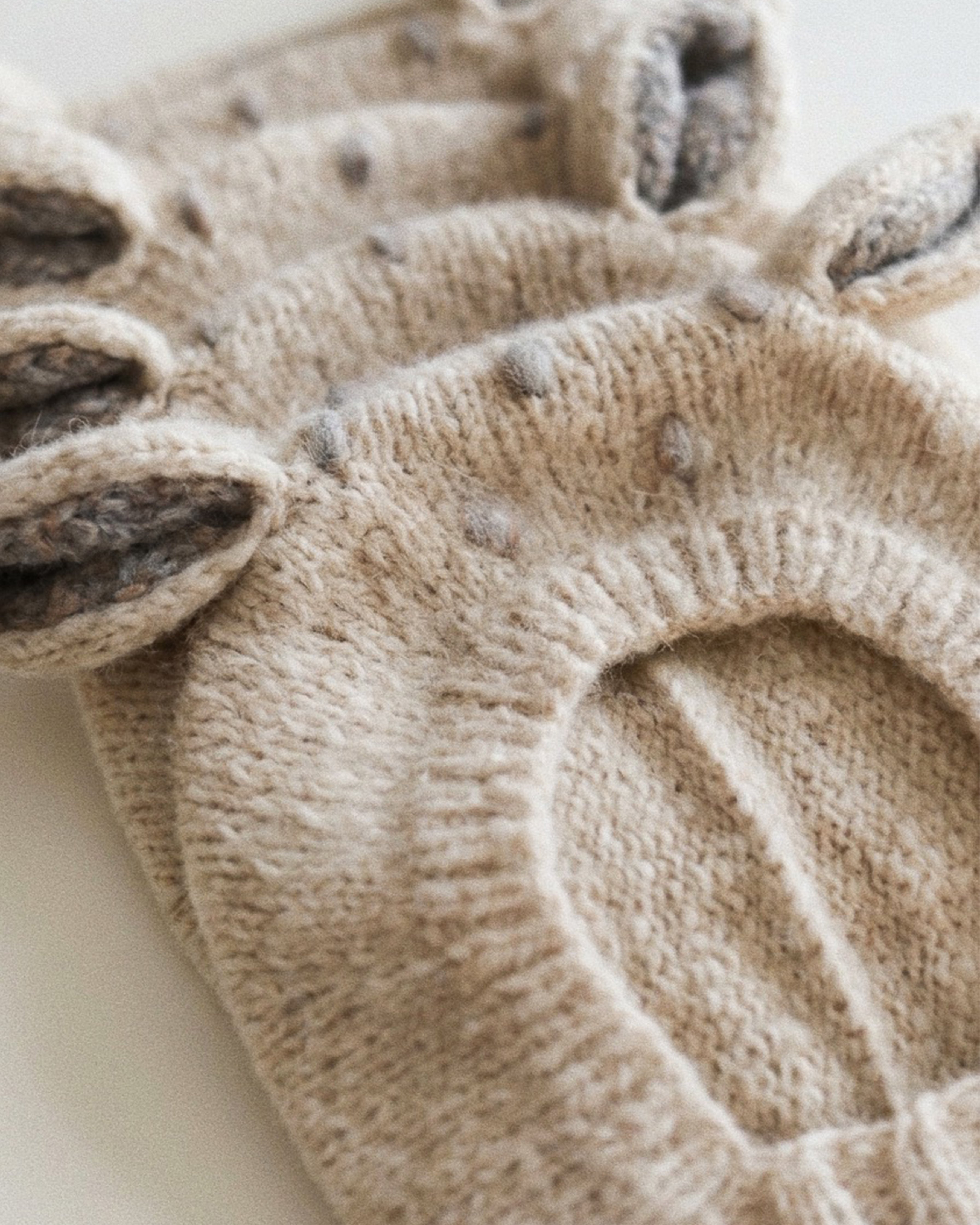 Close-up of a beige knit pet hood with ear flaps and small gray pom-pom accents, photographed on a light neutral background.