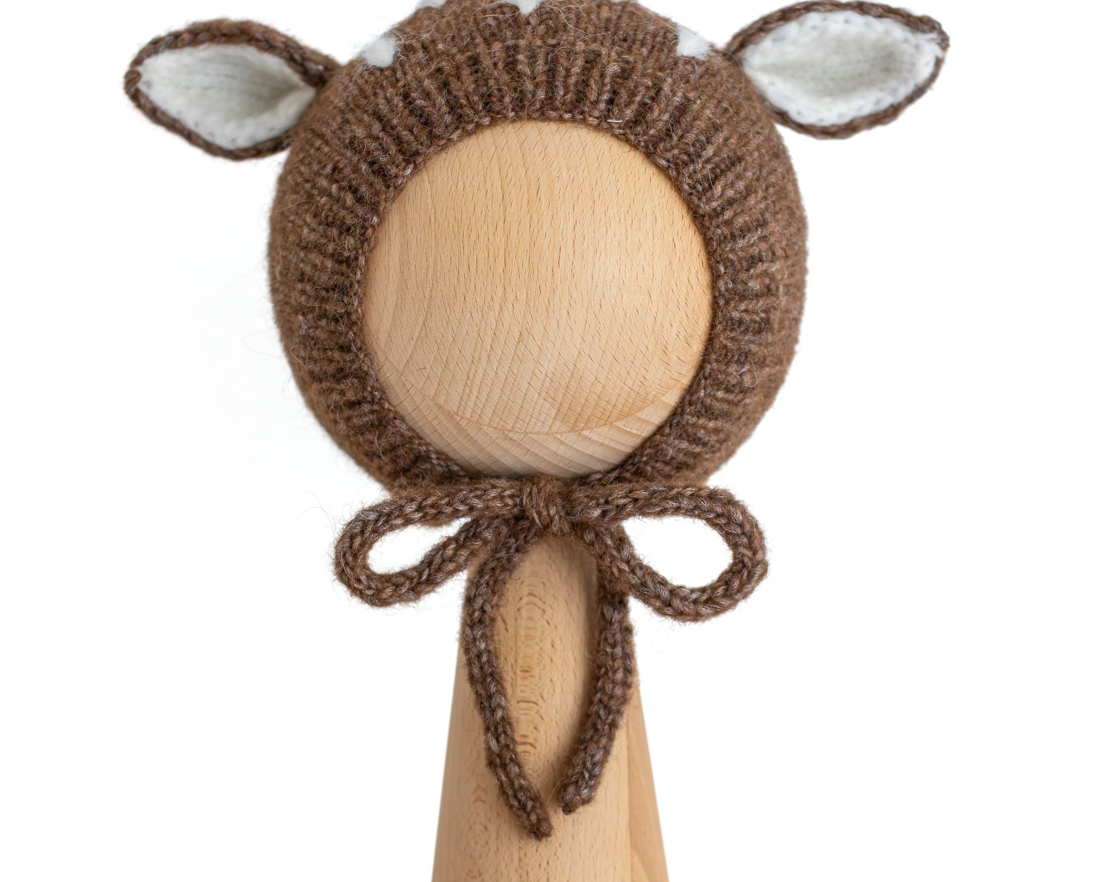 A brown knitted baby bonnet with floppy ears and a bow tie, displayed on a wooden mannequin head against a plain white background.