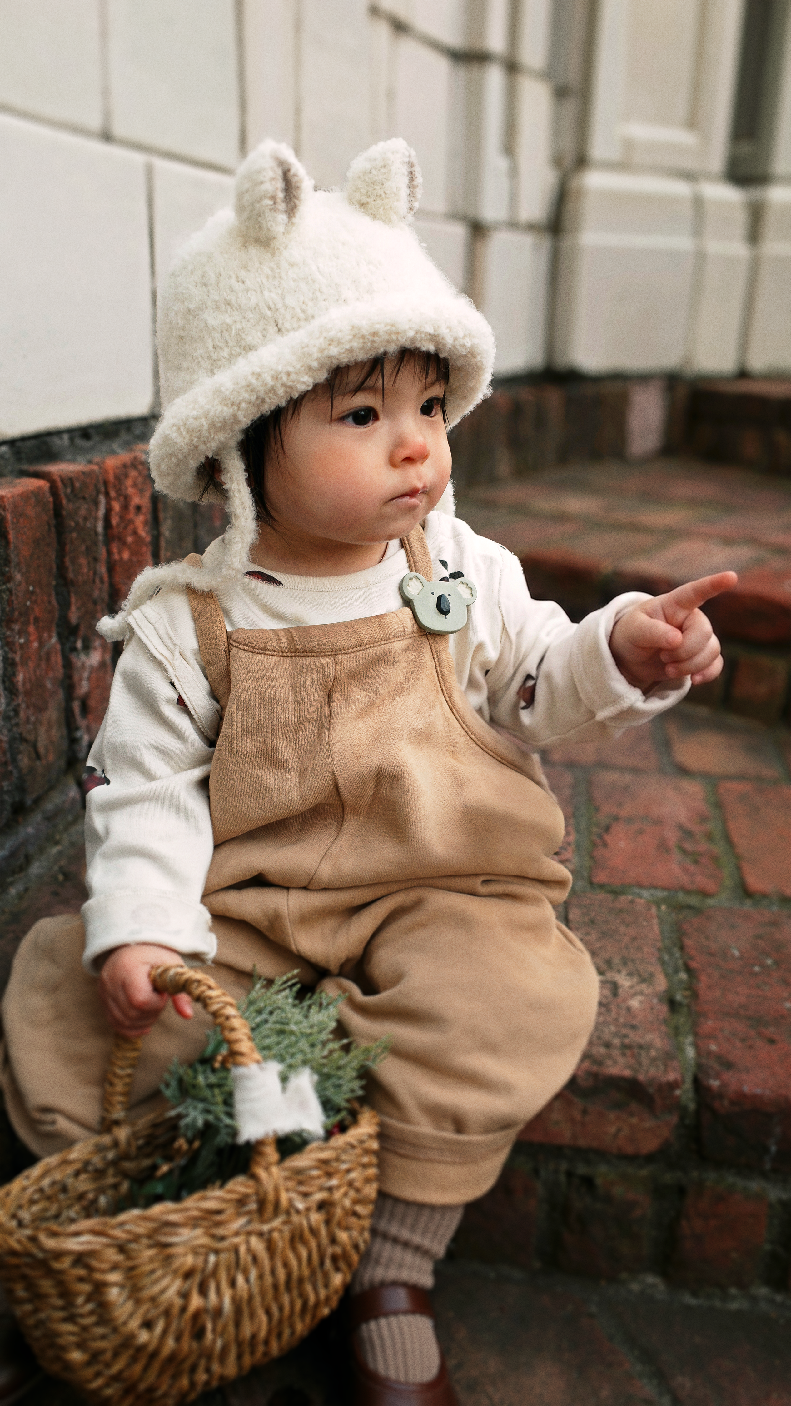 A baby wearing a cream fuzzy bunny hat, a white long-sleeve shirt, and tan corduroy overalls sits on red brick steps, holding a small woven basket with greenery.