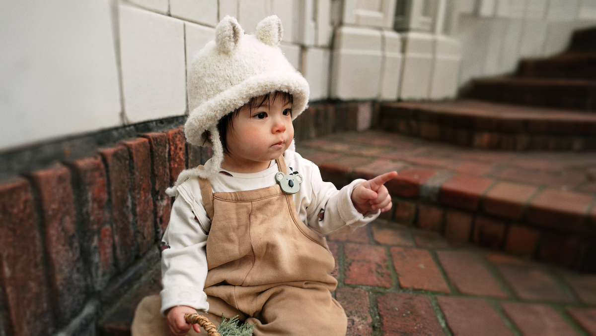 A baby wearing a cream-colored fuzzy bear hat with ear tips, a white long-sleeve shirt, and tan overalls, sitting on a brick bench in an outdoor setting.