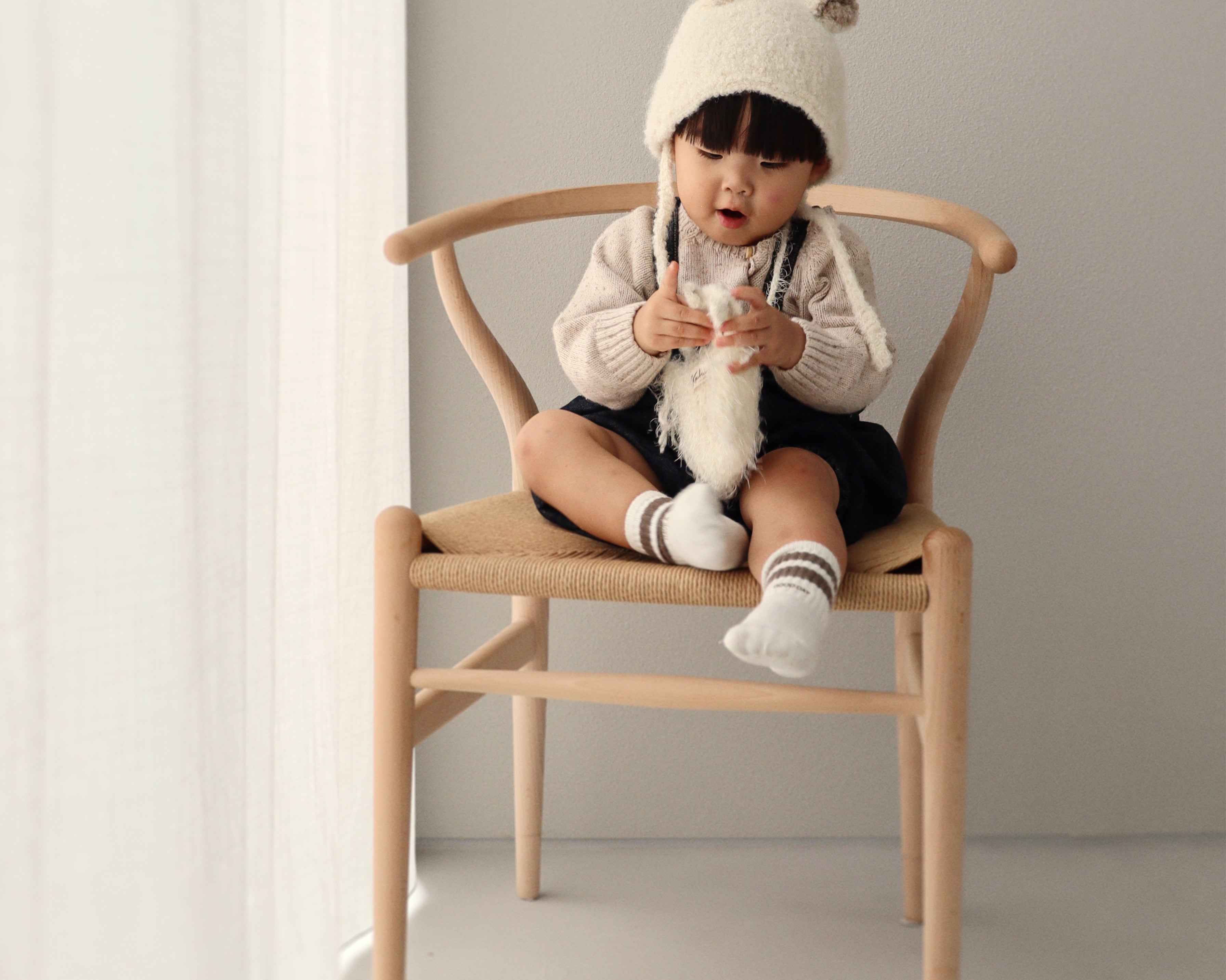 A child sitting on a wooden chair wearing the Bucket Bear Hat, a cozy knitted winter hat with bear ears.