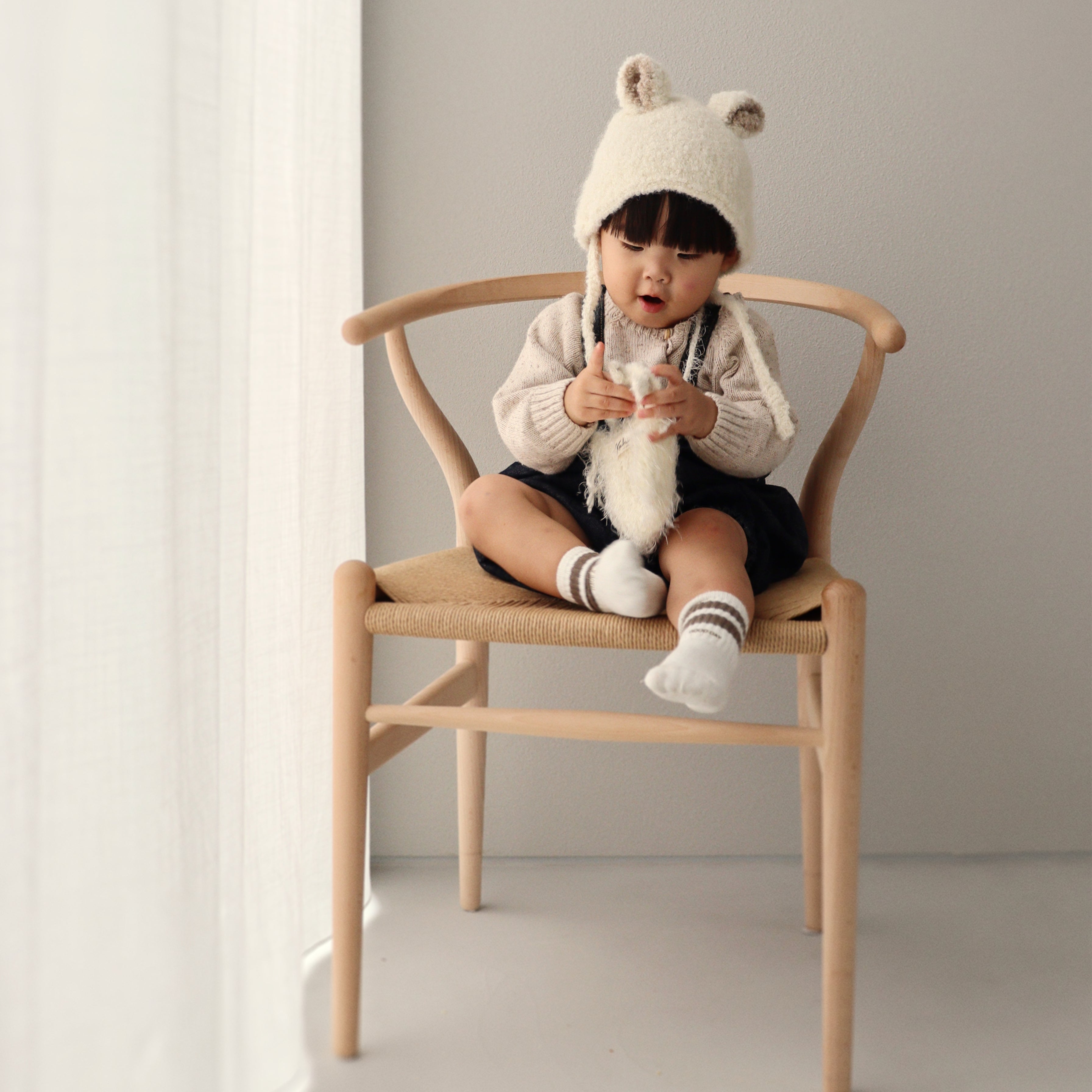 A child sitting on a wooden chair wearing the Bucket Bear Hat, a cozy knitted winter hat with bear ears.