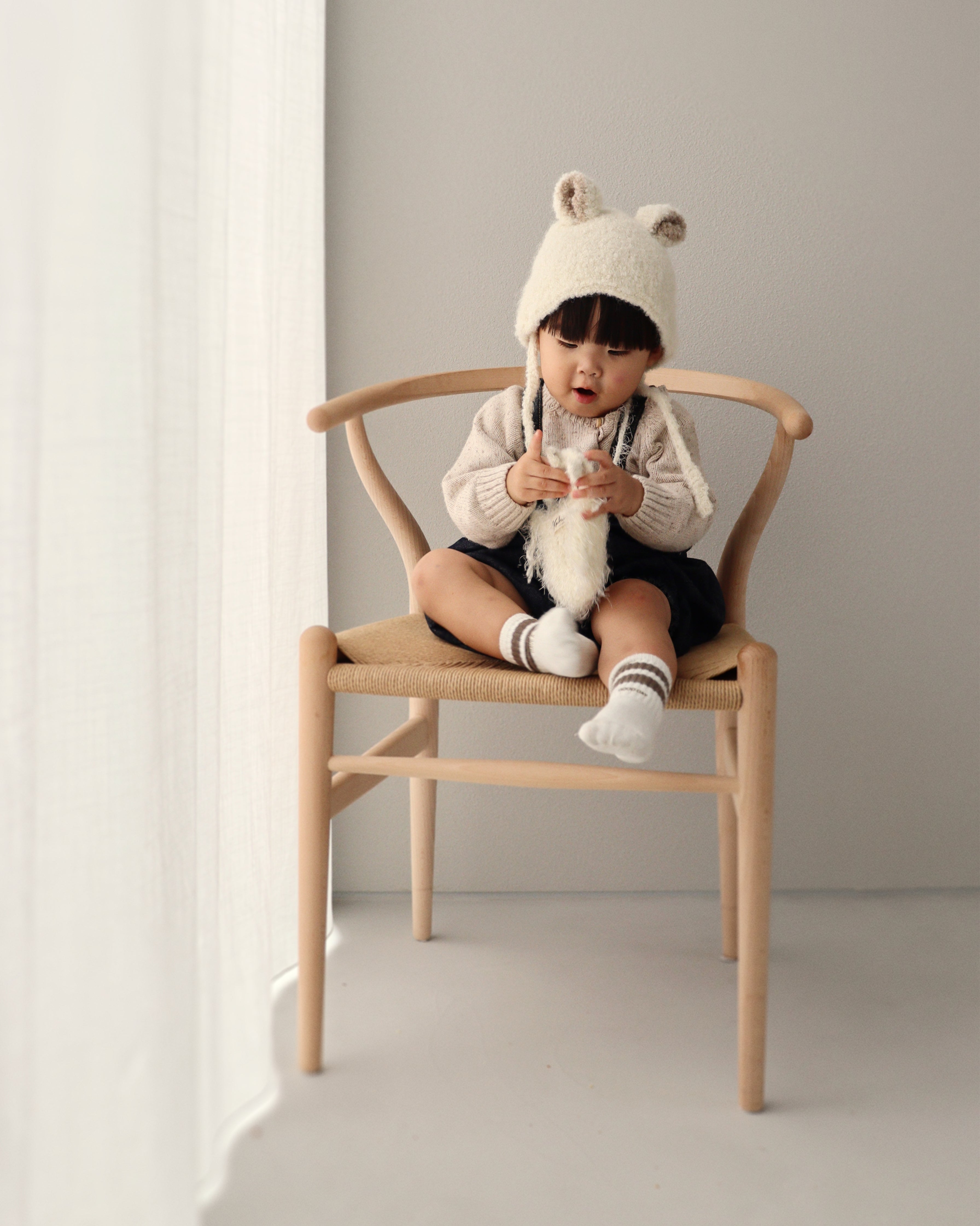 A child sitting on a wooden chair wearing the Bucket Bear Hat, a cozy knitted winter hat with bear ears.