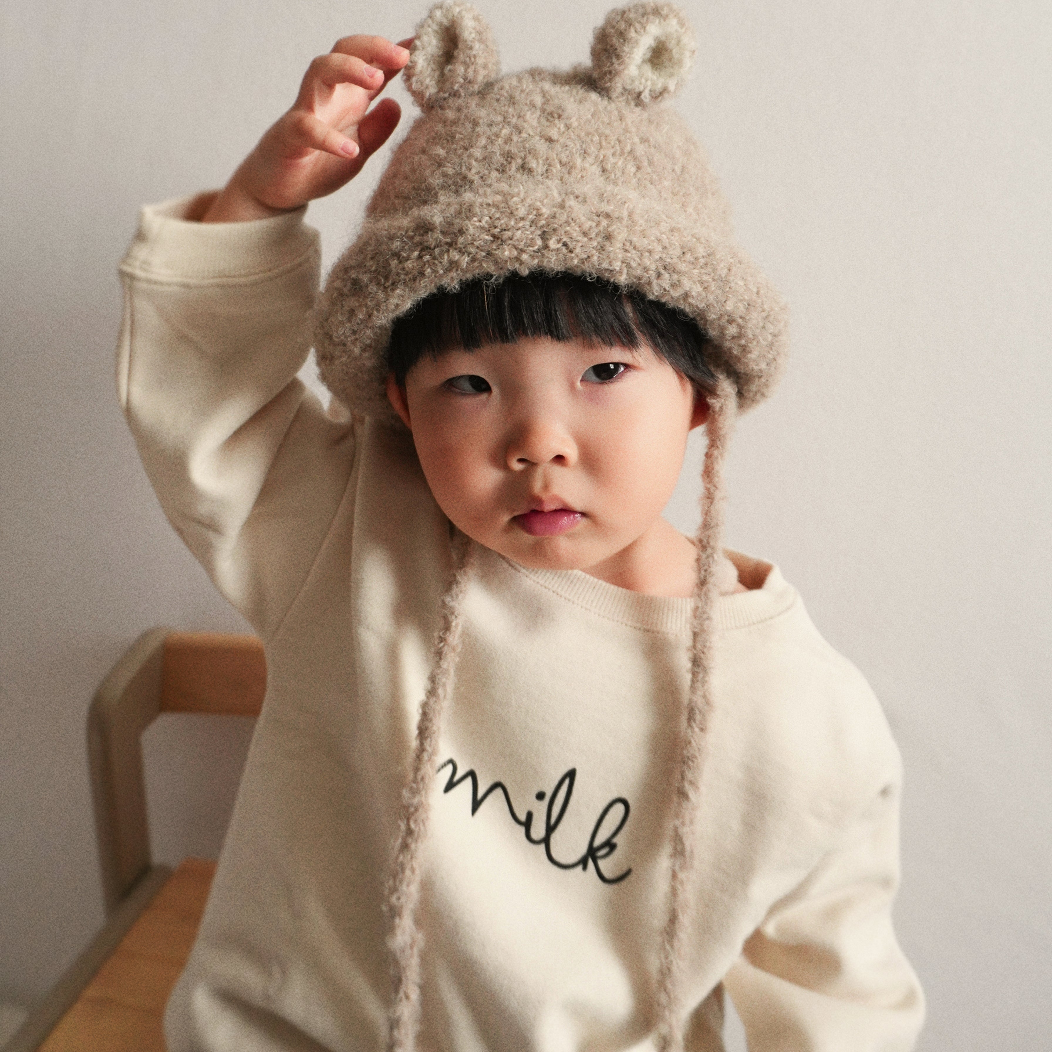 A toddler playfully holding the ear of the Bucket Bear Hat in beige, a knitted bucket hat with bear ears from Gang of Kids.