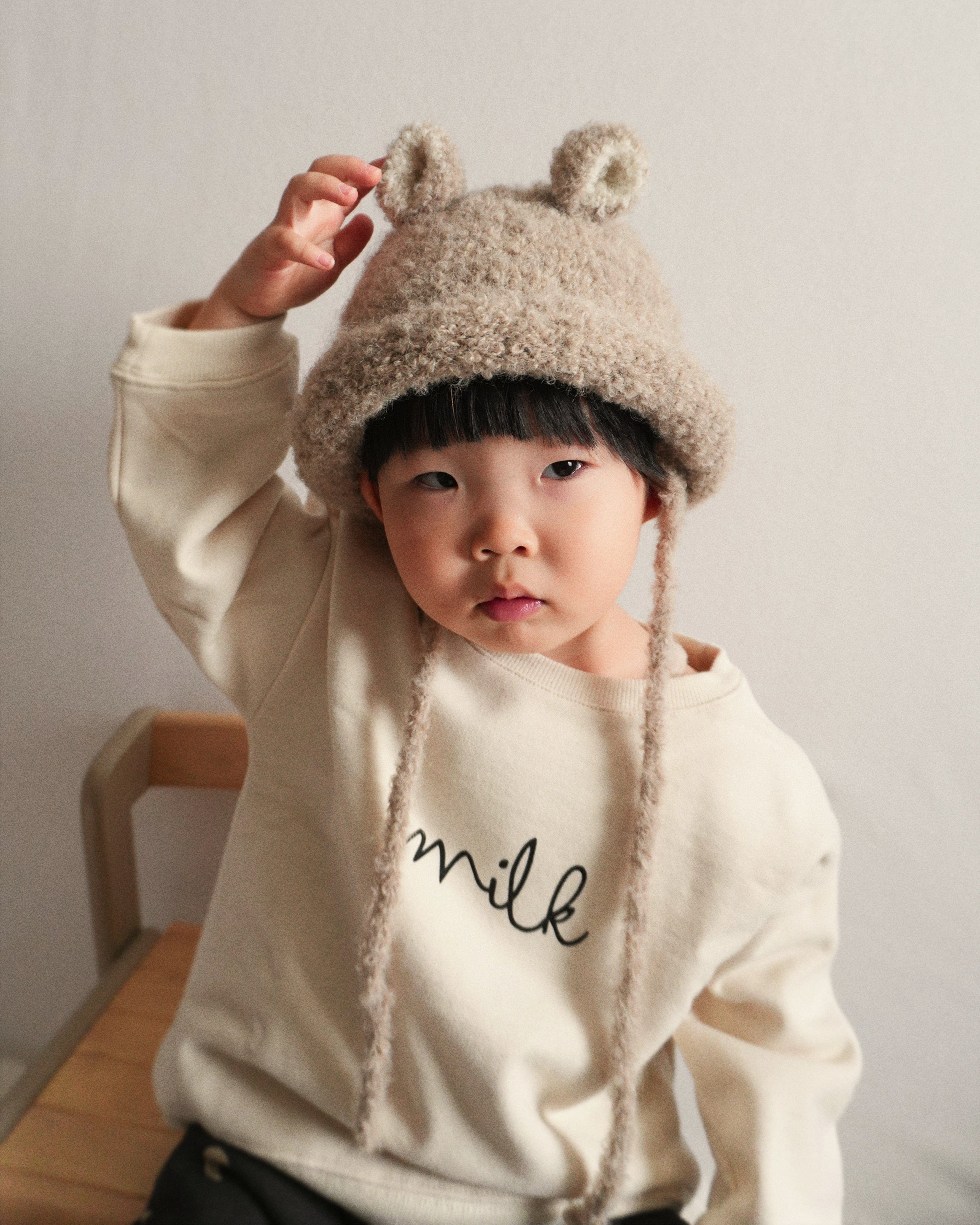 A toddler playfully holding the ear of the Bucket Bear Hat in beige, a knitted bucket hat with bear ears from Gang of Kids.