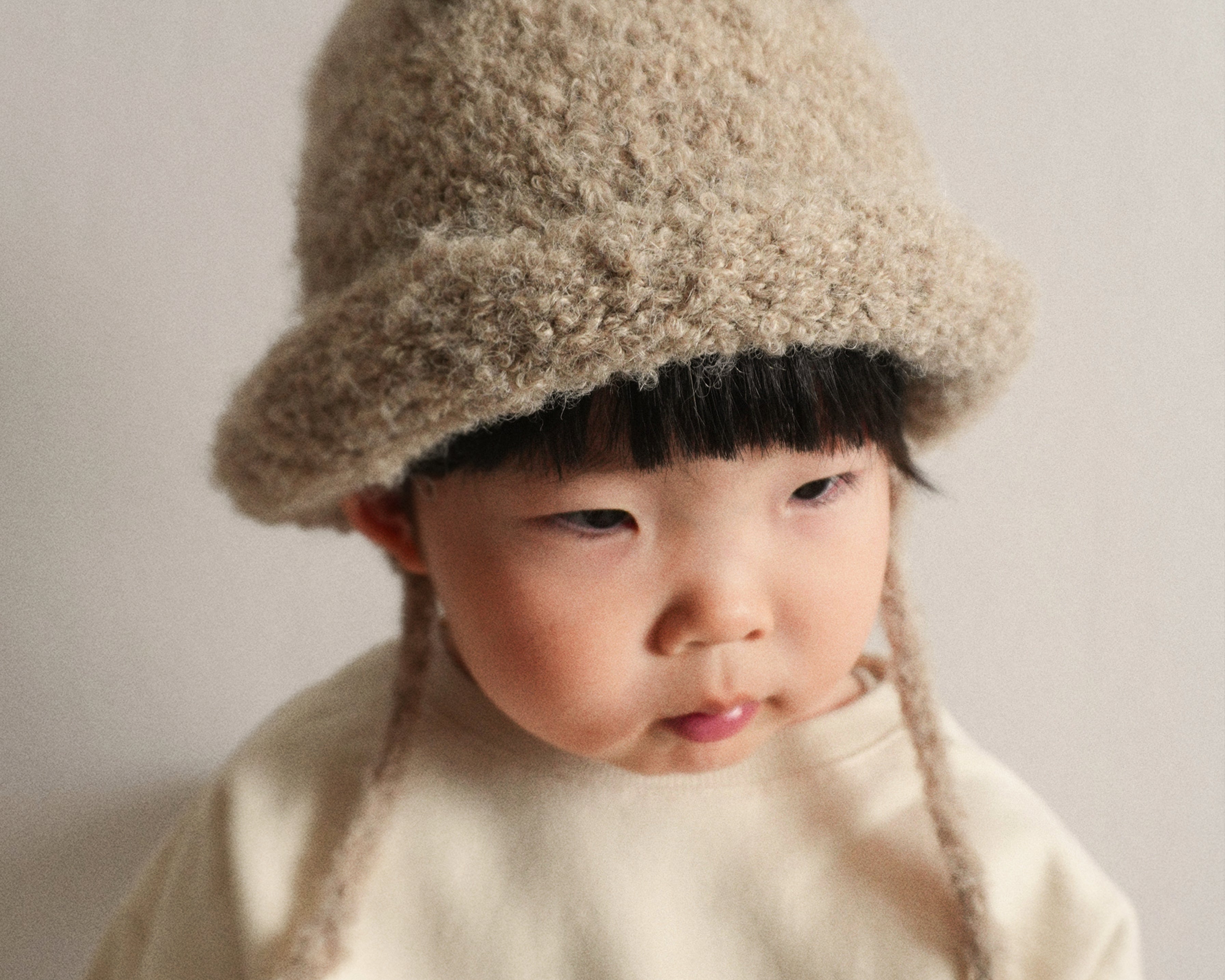 A portrait of a child wearing the Bucket Bear Hat in beige, a knitted bucket hat with bear ears, from the Gang of Kids winter collection.