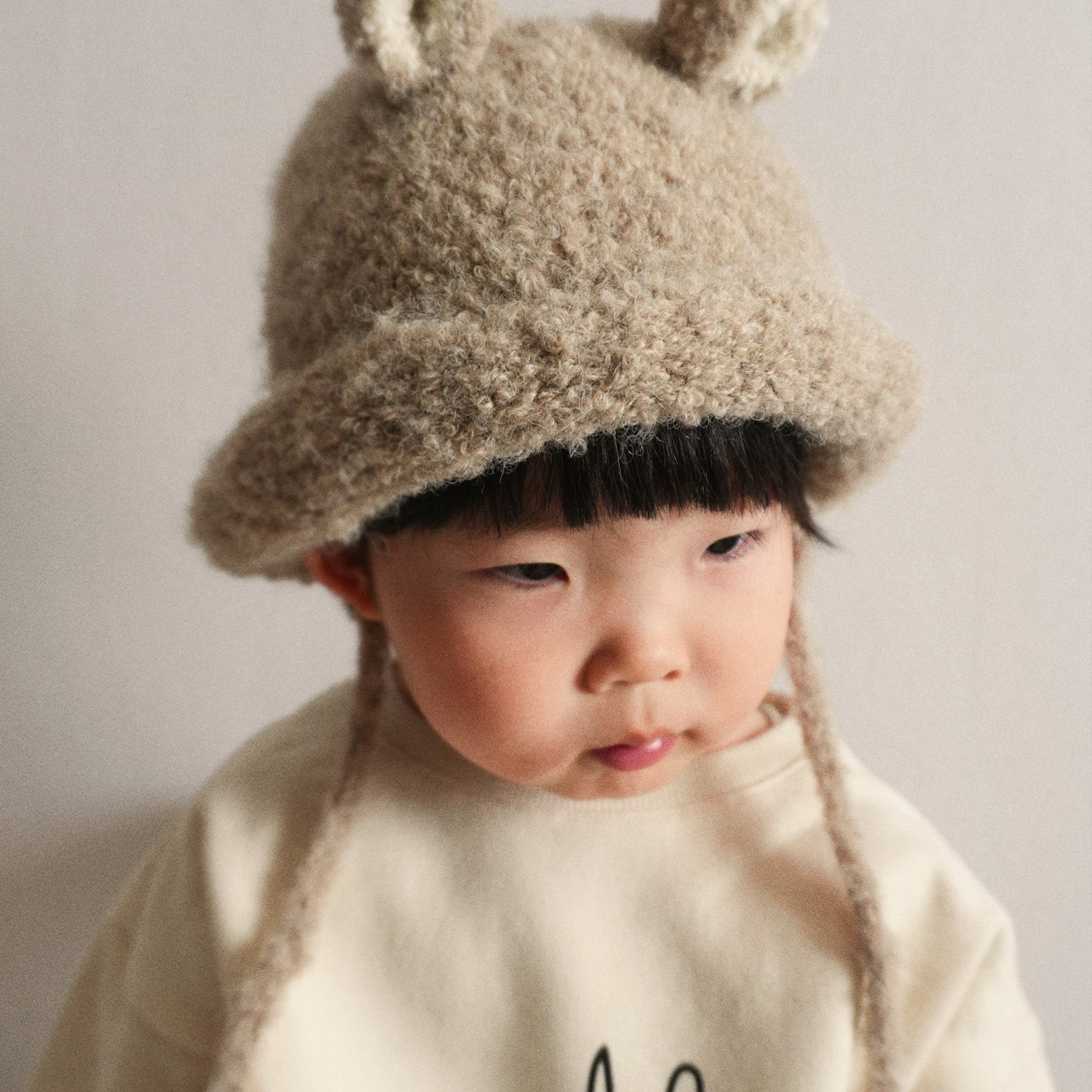 A portrait of a child wearing the Bucket Bear Hat in beige, a knitted bucket hat with bear ears, from the Gang of Kids winter collection.