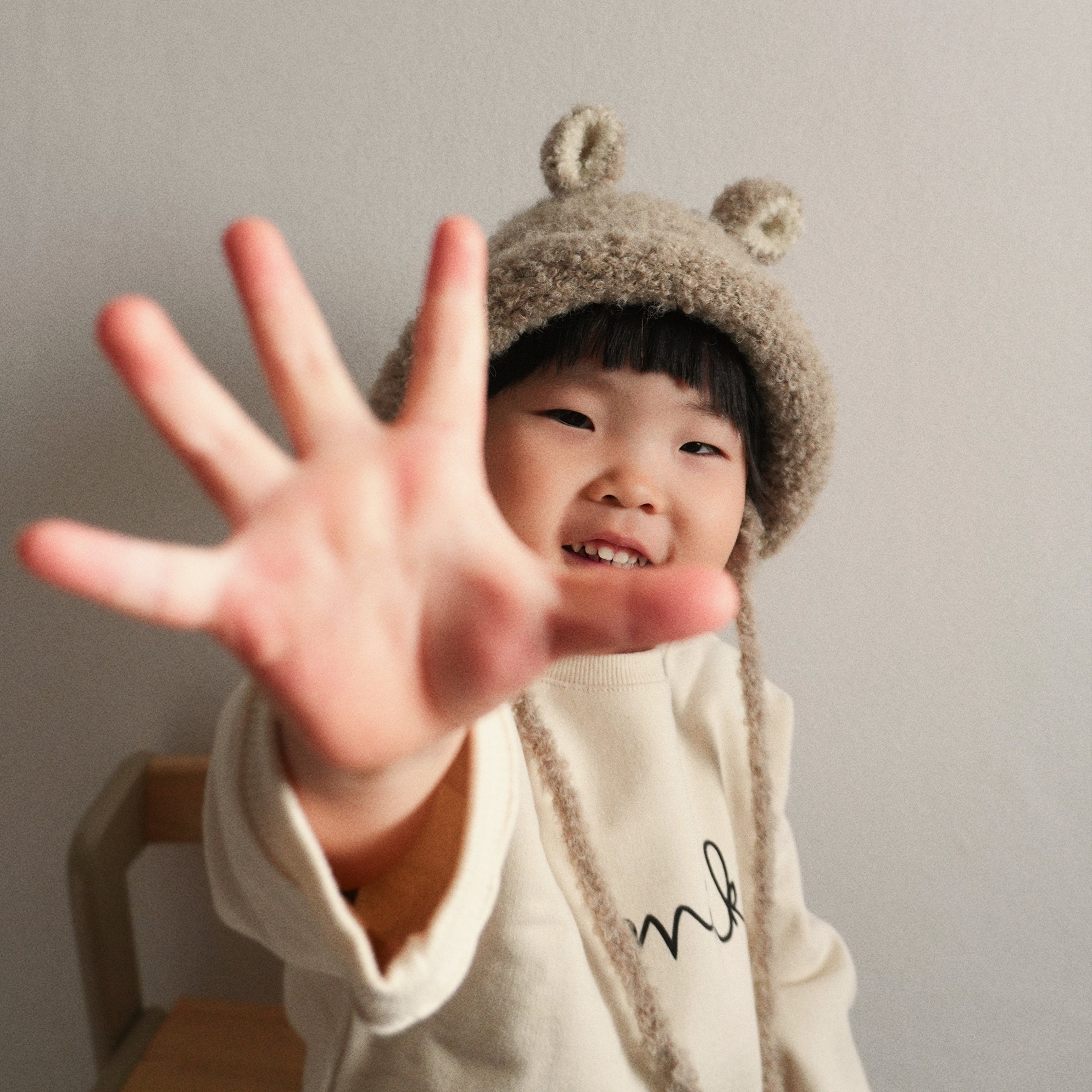 A smiling child reaching toward the camera while wearing the Bucket Bear Hat in beige, a cozy knitted winter hat with bear ears.