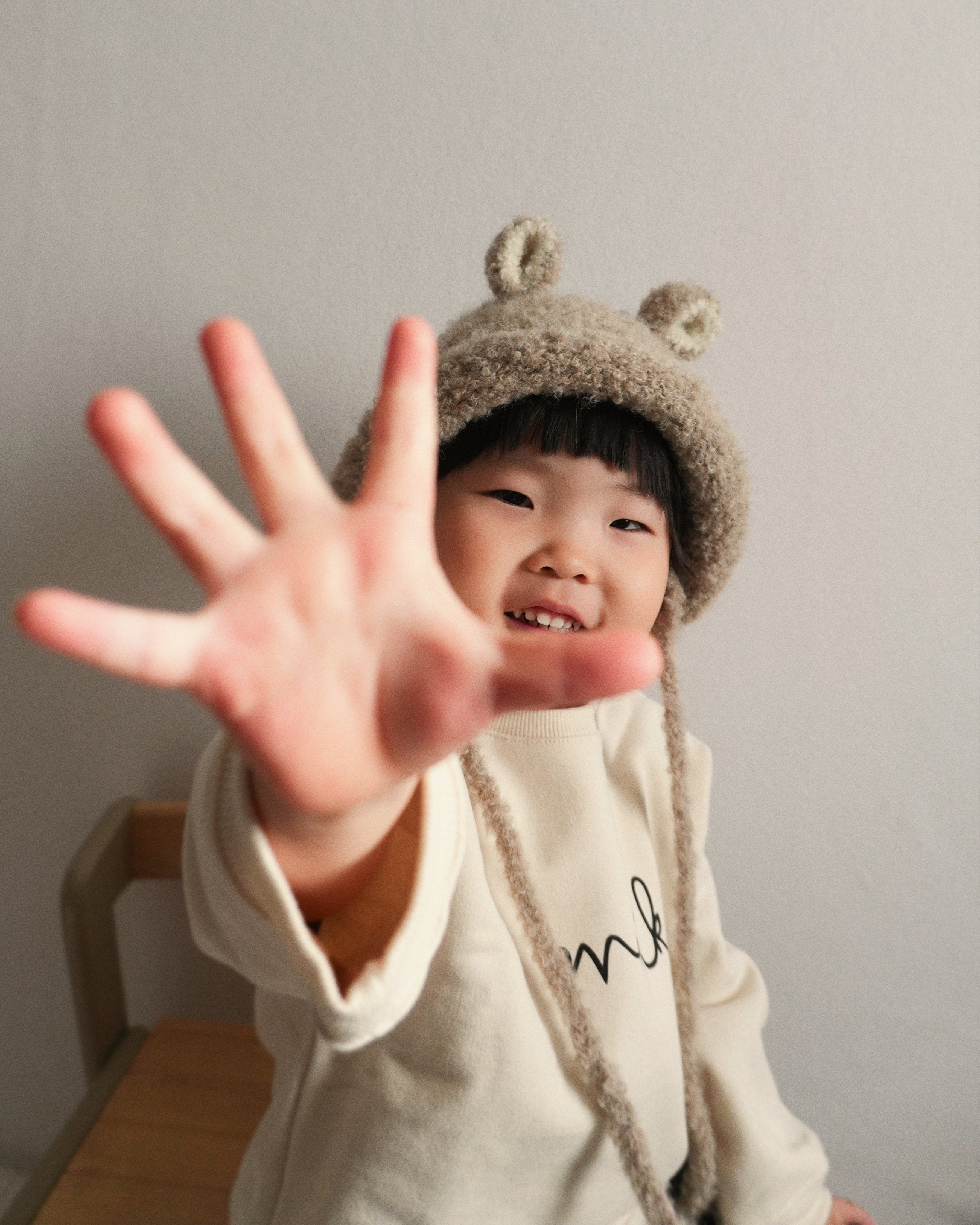 A smiling child reaching toward the camera while wearing the Bucket Bear Hat in beige, a cozy knitted winter hat with bear ears.