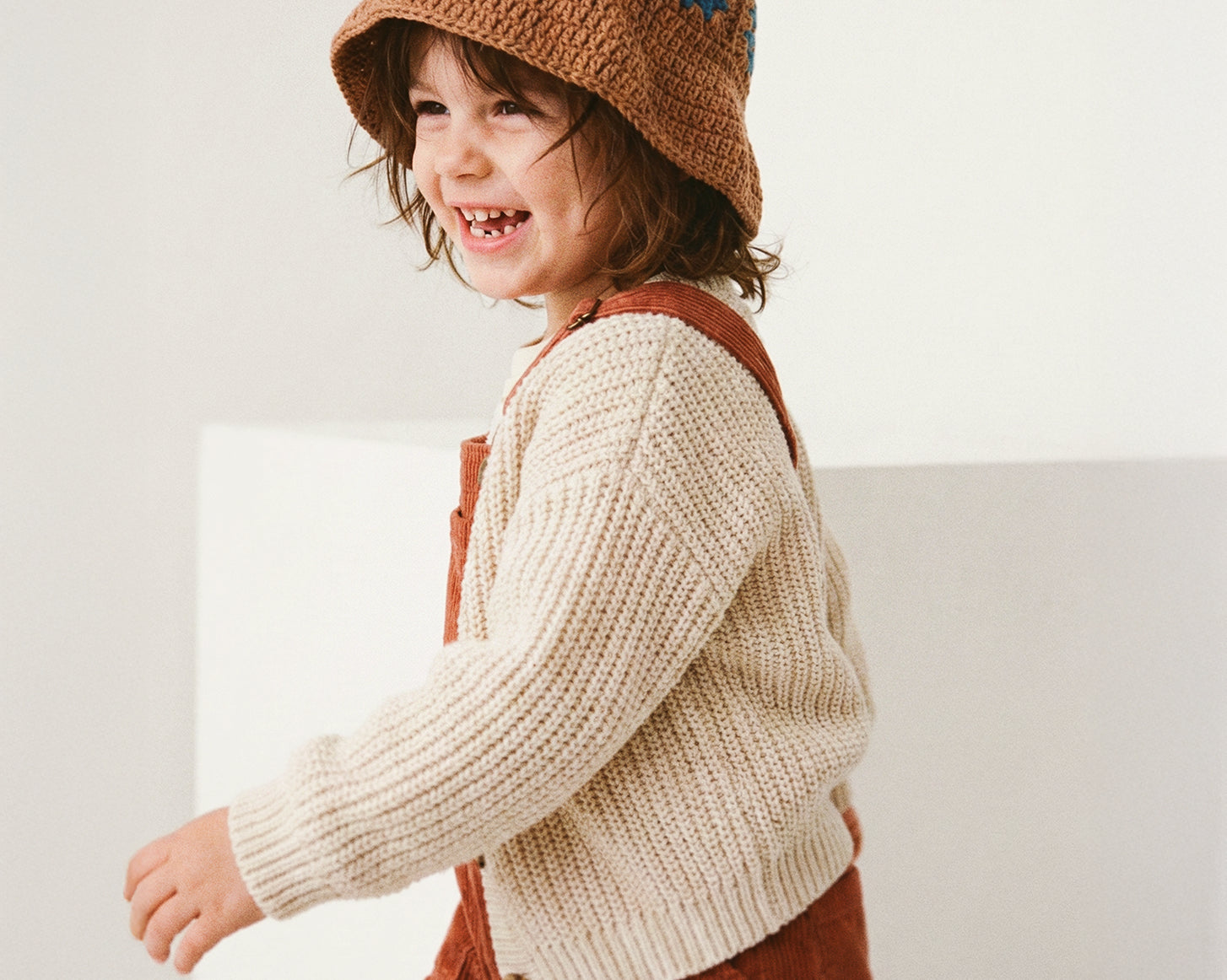 Uśmiechnięte dziecko biegnące w brązowym szydełkowym kapeluszu Daisy Boho Earth Teal w beżowym swetrze i rudych ogrodniczkach | Smiling toddler running in Boho Earth Teal Daisy crocheted hat with beige sweater and rust dungarees