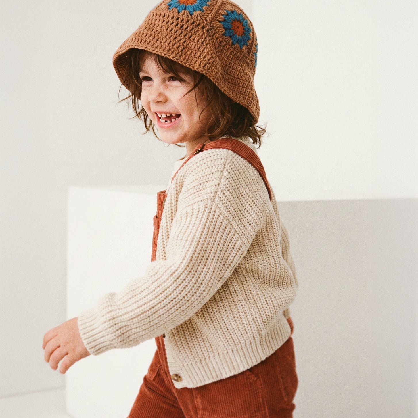 Uśmiechnięte dziecko biegnące w brązowym szydełkowym kapeluszu Daisy Boho Earth Teal w beżowym swetrze i rudych ogrodniczkach | Smiling toddler running in Boho Earth Teal Daisy crocheted hat with beige sweater and rust dungarees
