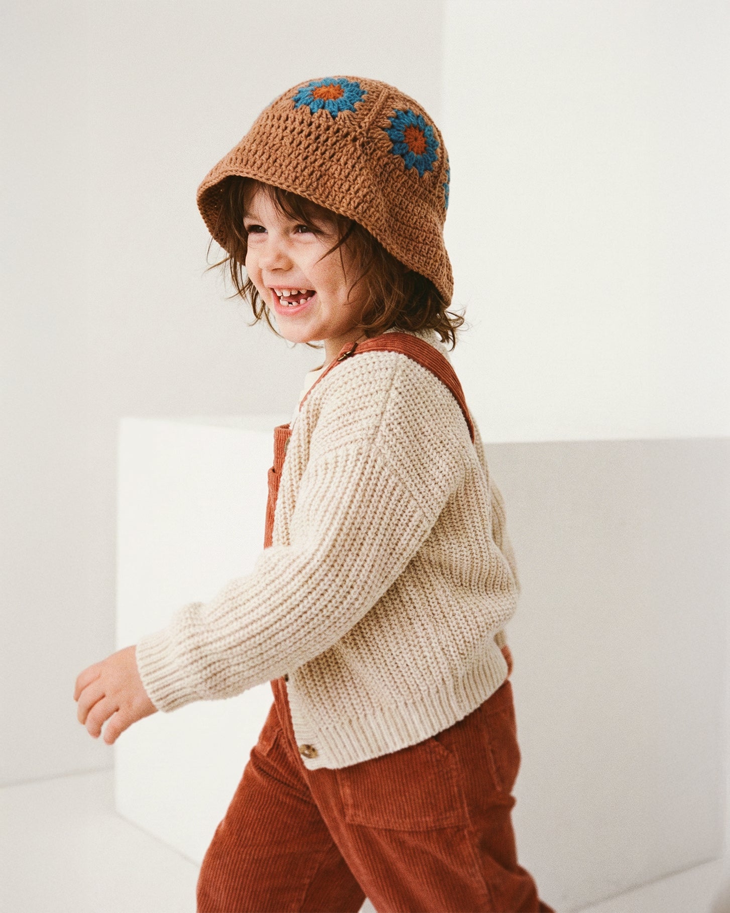 Uśmiechnięte dziecko biegnące w brązowym szydełkowym kapeluszu Daisy Boho Earth Teal w beżowym swetrze i rudych ogrodniczkach | Smiling toddler running in Boho Earth Teal Daisy crocheted hat with beige sweater and rust dungarees