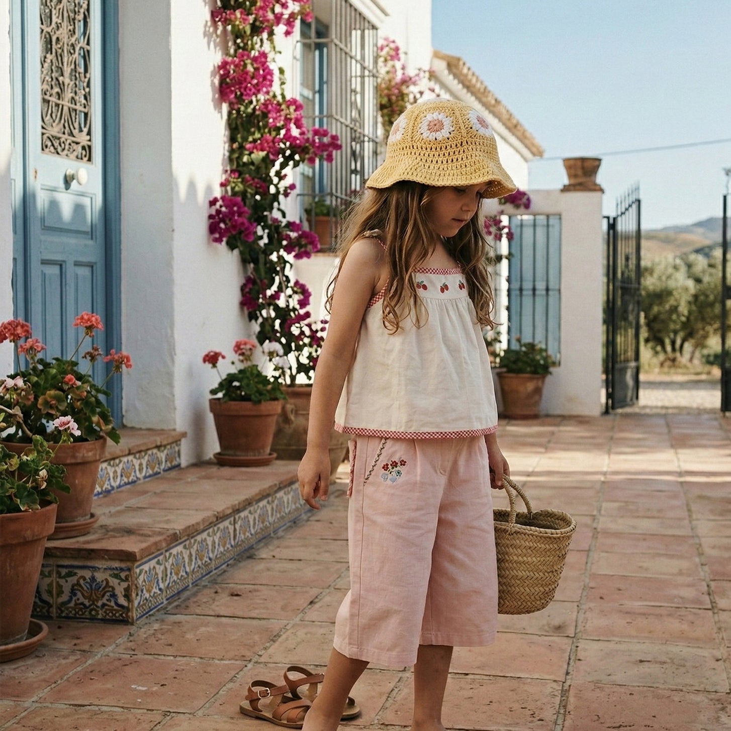 Dziecko w żółtym szydełkowym kapeluszu dziecięcym Daisy z koszykiem przy śródziemnomorskiej fasadzie | Child wearing yellow Daisy crochet sun hat with basket at Mediterranean house