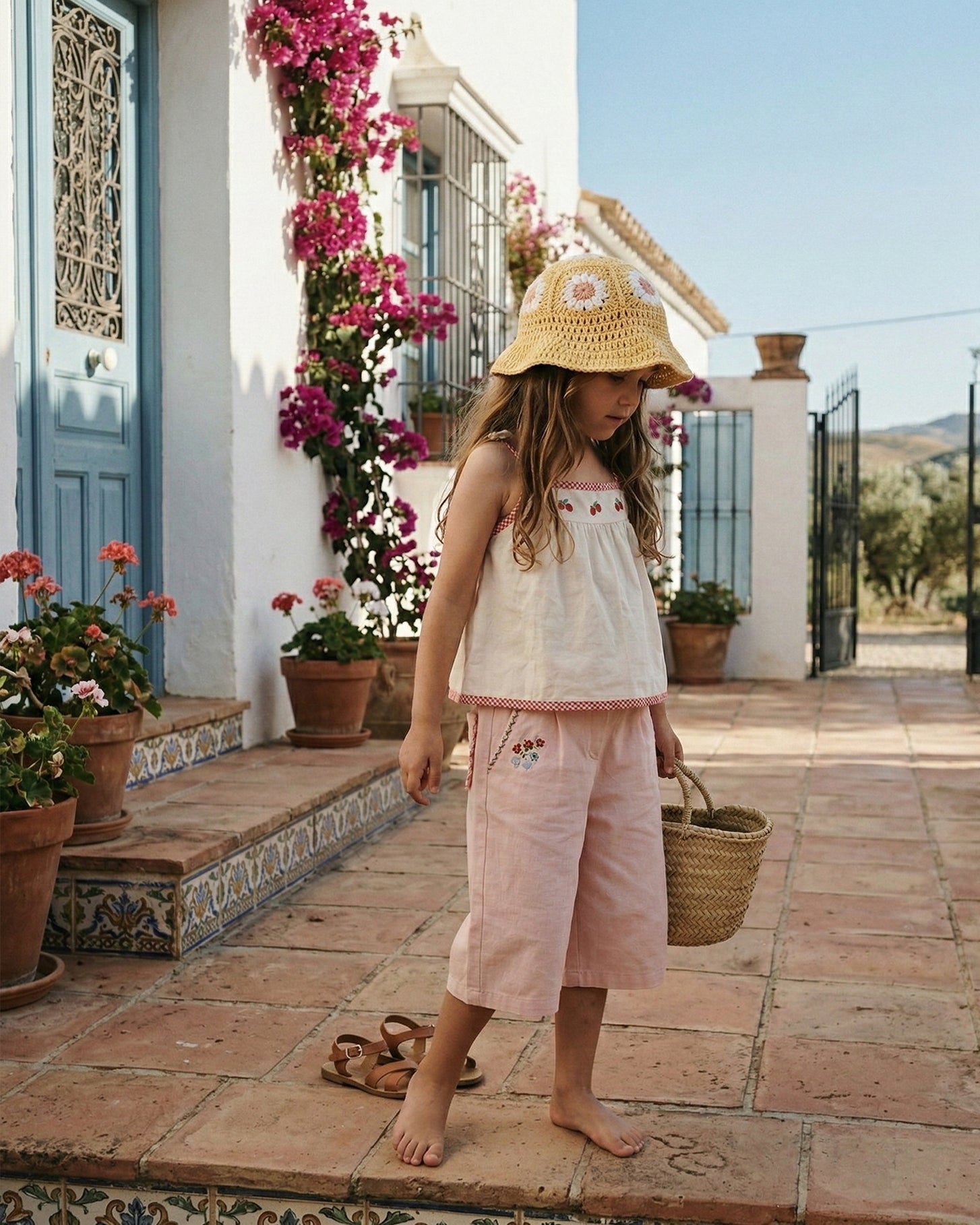 Dziecko w żółtym szydełkowym kapeluszu dziecięcym Daisy z koszykiem przy śródziemnomorskiej fasadzie | Child wearing yellow Daisy crochet sun hat with basket at Mediterranean house