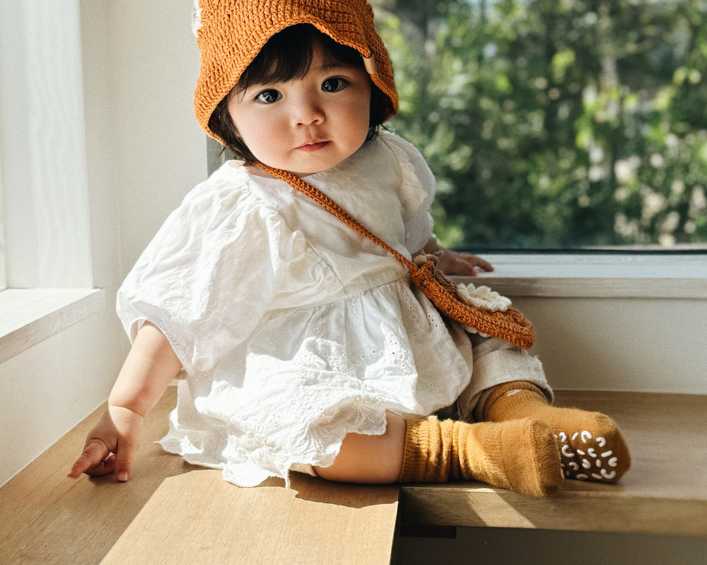Niemowlę w bursztynowym szydełkowym kapeluszu Daisy Honey Amber siedzące przy oknie w białej sukience | Baby wearing honey amber Daisy crocheted hat sitting by window in white dress