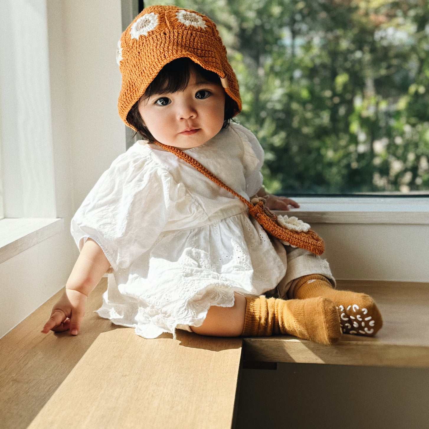 Niemowlę w bursztynowym szydełkowym kapeluszu Daisy Honey Amber siedzące przy oknie w białej sukience | Baby wearing honey amber Daisy crocheted hat sitting by window in white dress