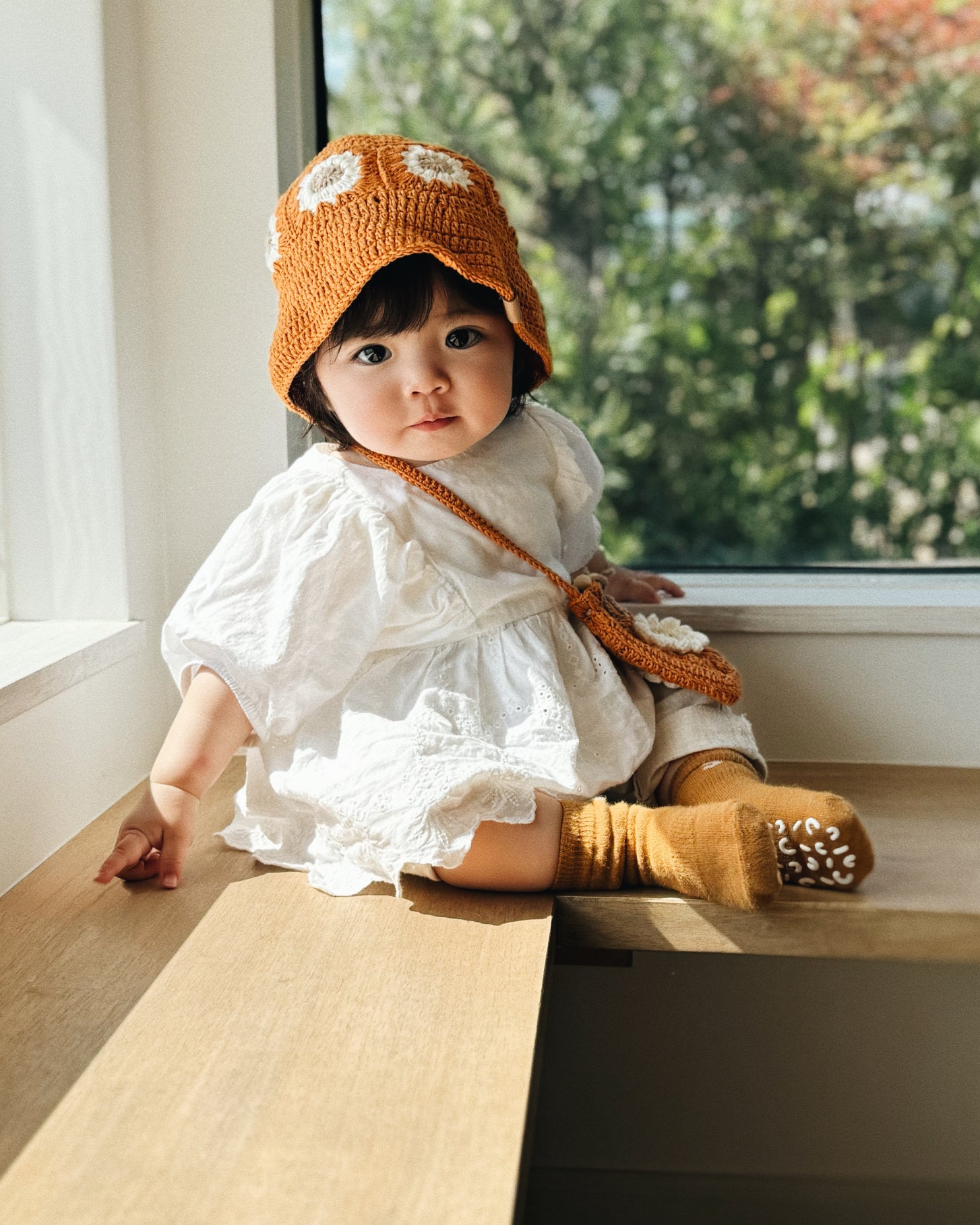 Niemowlę w bursztynowym szydełkowym kapeluszu Daisy Honey Amber siedzące przy oknie w białej sukience | Baby wearing honey amber Daisy crocheted hat sitting by window in white dress