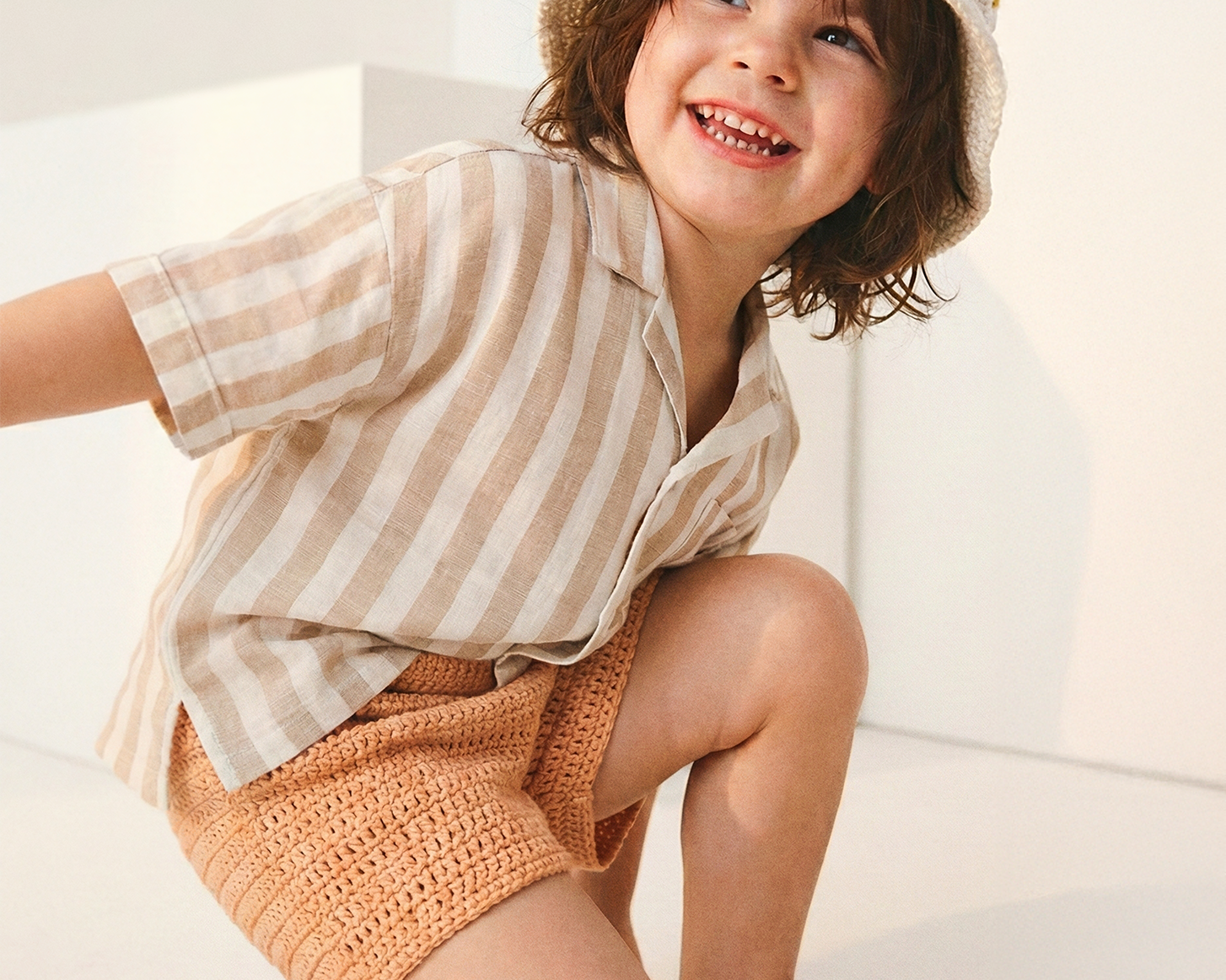 Uśmiechnięte dziecko w kremowym szydełkowym kapeluszu Daisy Vanilla Cream i pasiastej koszuli | Smiling toddler wearing Daisy Vanilla Cream crocheted sun hat with striped linen shirt