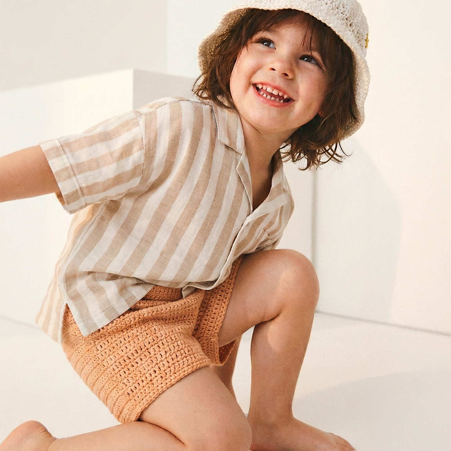 Uśmiechnięte dziecko w kremowym szydełkowym kapeluszu Daisy Vanilla Cream i pasiastej koszuli | Smiling toddler wearing Daisy Vanilla Cream crocheted sun hat with striped linen shirt