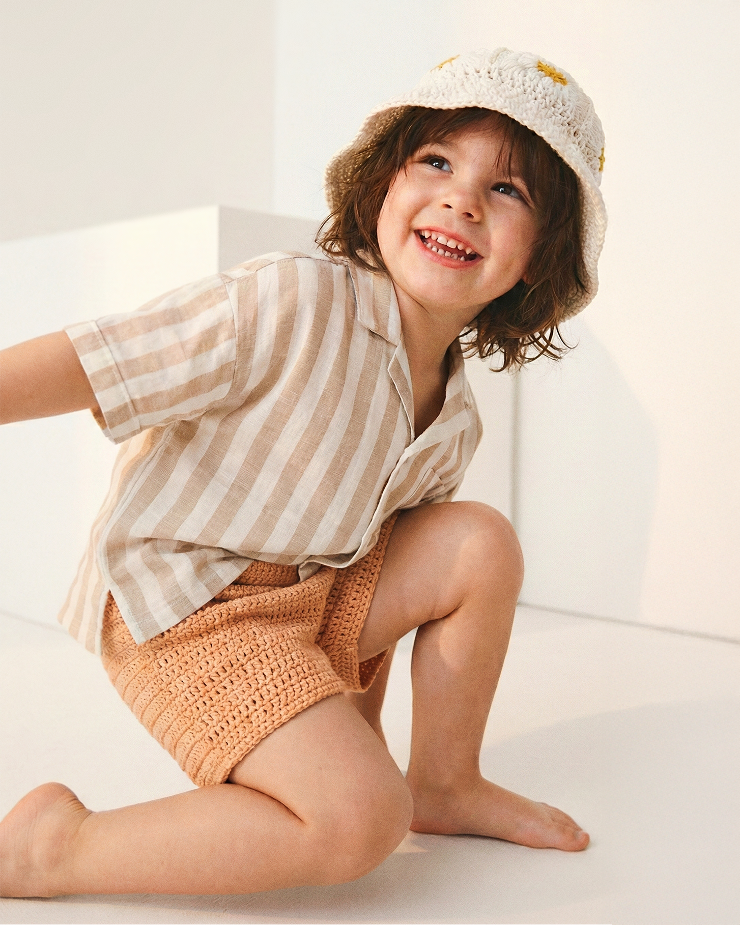 Uśmiechnięte dziecko w kremowym szydełkowym kapeluszu Daisy Vanilla Cream i pasiastej koszuli | Smiling toddler wearing Daisy Vanilla Cream crocheted sun hat with striped linen shirt