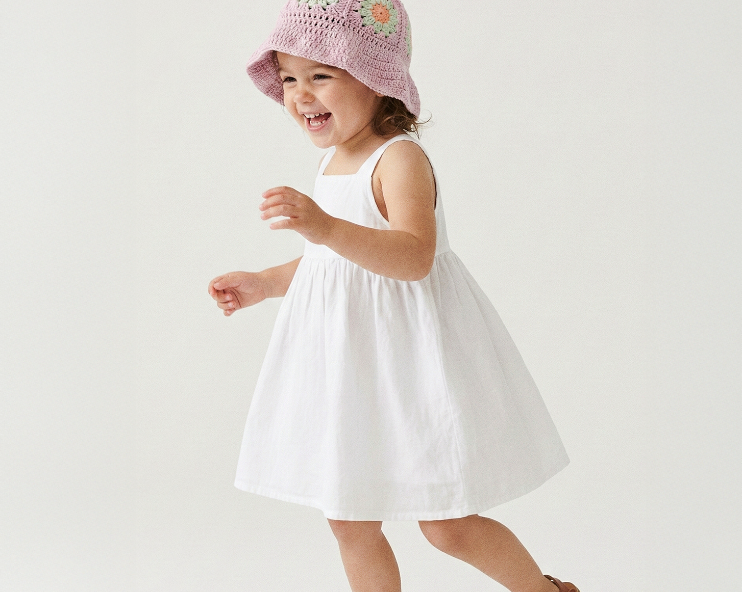 Uśmiechnięta dziewczynka tańcząca w pastelowo-fioletowym szydełkowym kapeluszu Daisy Wild Berry Sorbet i białej sukience | Smiling toddler girl dancing in pastel purple Wild Berry Sorbet Daisy crocheted sun hat and white dress