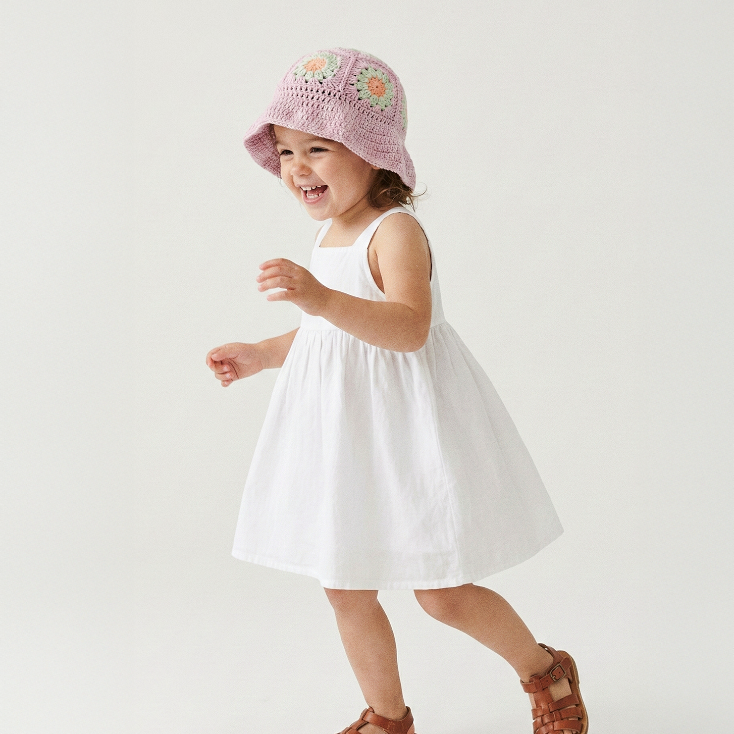 Uśmiechnięta dziewczynka tańcząca w pastelowo-fioletowym szydełkowym kapeluszu Daisy Wild Berry Sorbet i białej sukience | Smiling toddler girl dancing in pastel purple Wild Berry Sorbet Daisy crocheted sun hat and white dress