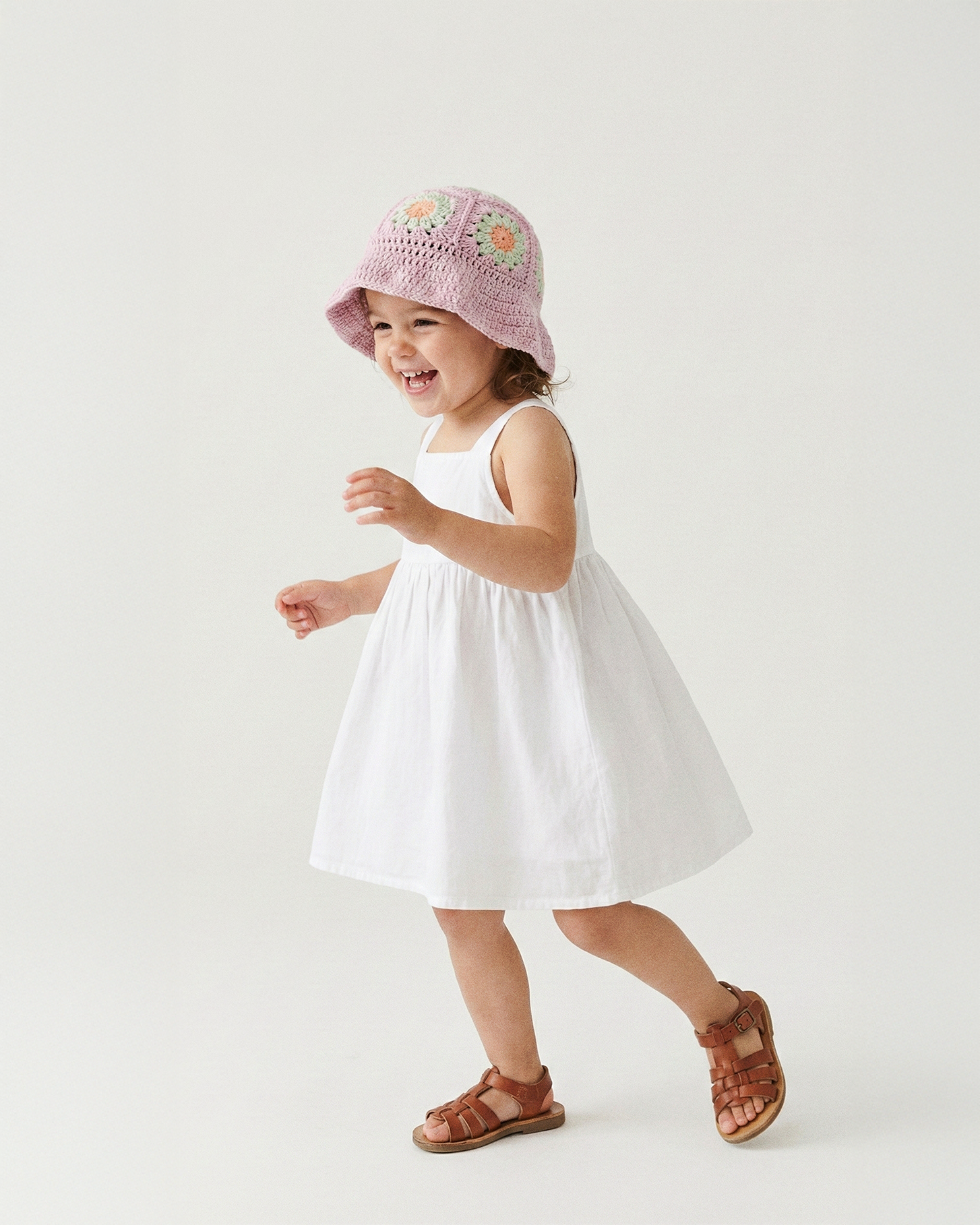 Uśmiechnięta dziewczynka tańcząca w pastelowo-fioletowym szydełkowym kapeluszu Daisy Wild Berry Sorbet i białej sukience | Smiling toddler girl dancing in pastel purple Wild Berry Sorbet Daisy crocheted sun hat and white dress
