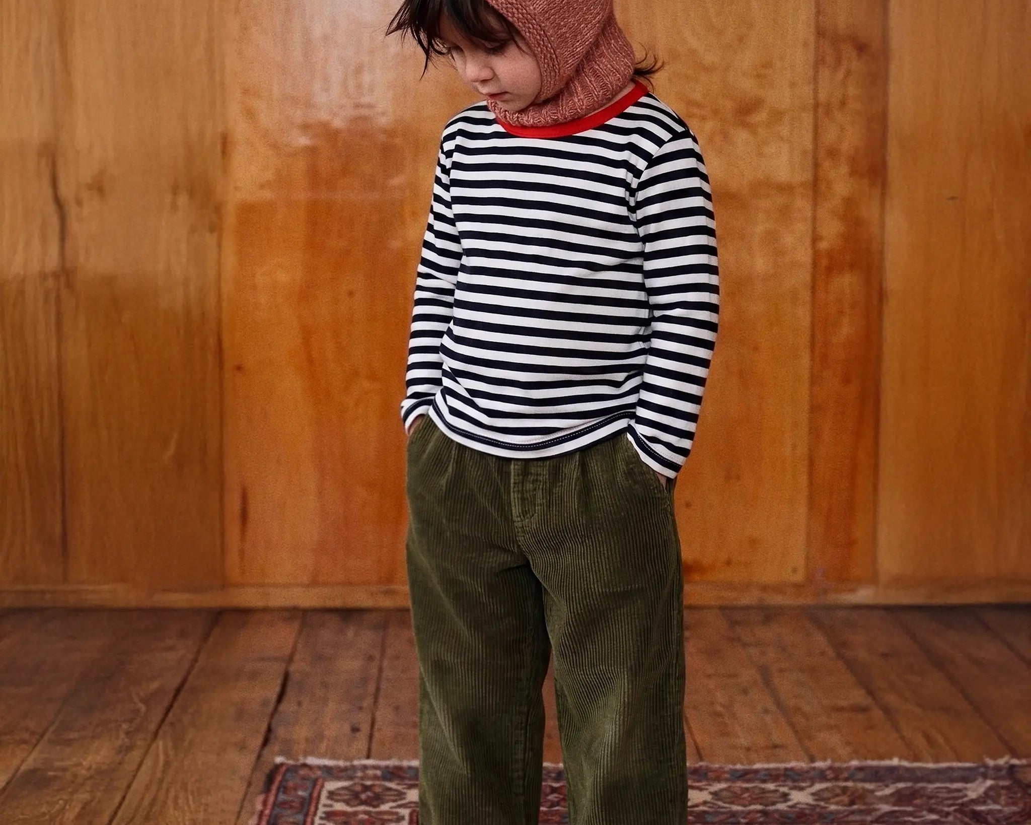 A toddler standing barefoot on a patterned rug in front of a wooden wall, wearing a black and white striped long-sleeve shirt, olive green corduroy pants, and a dusty rose knit hood with bear ears.