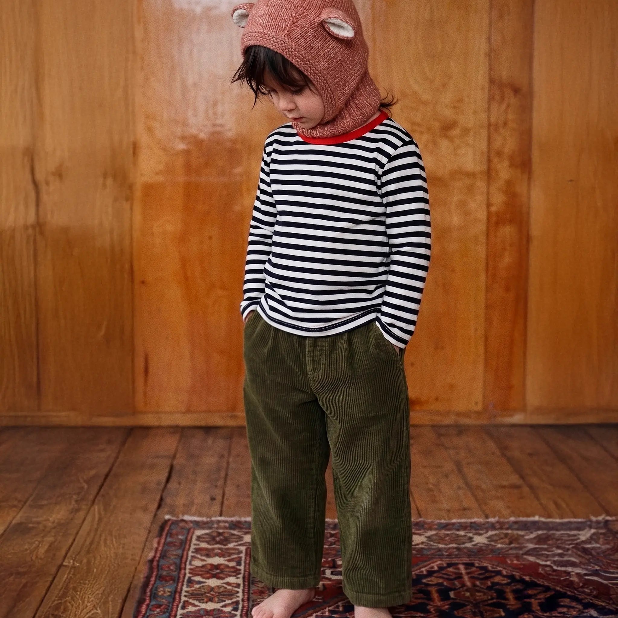 A toddler standing barefoot on a patterned rug in front of a wooden wall, wearing a black and white striped long-sleeve shirt, olive green corduroy pants, and a dusty rose knit hood with bear ears.