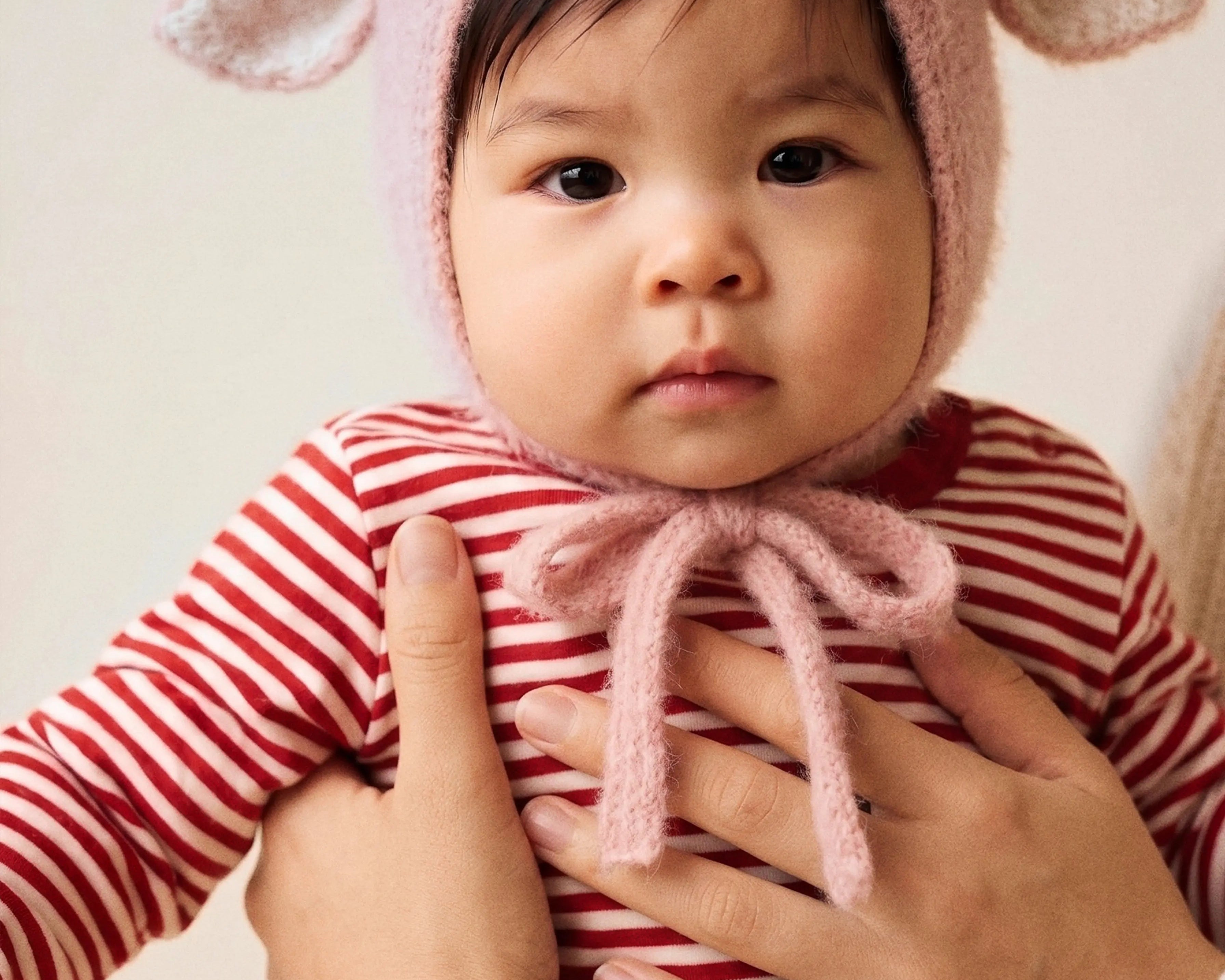 A baby held in a parent's arms wearing the Fairy Hat in Baby Pink, with the soft knitted ties loosely tied under the chin.