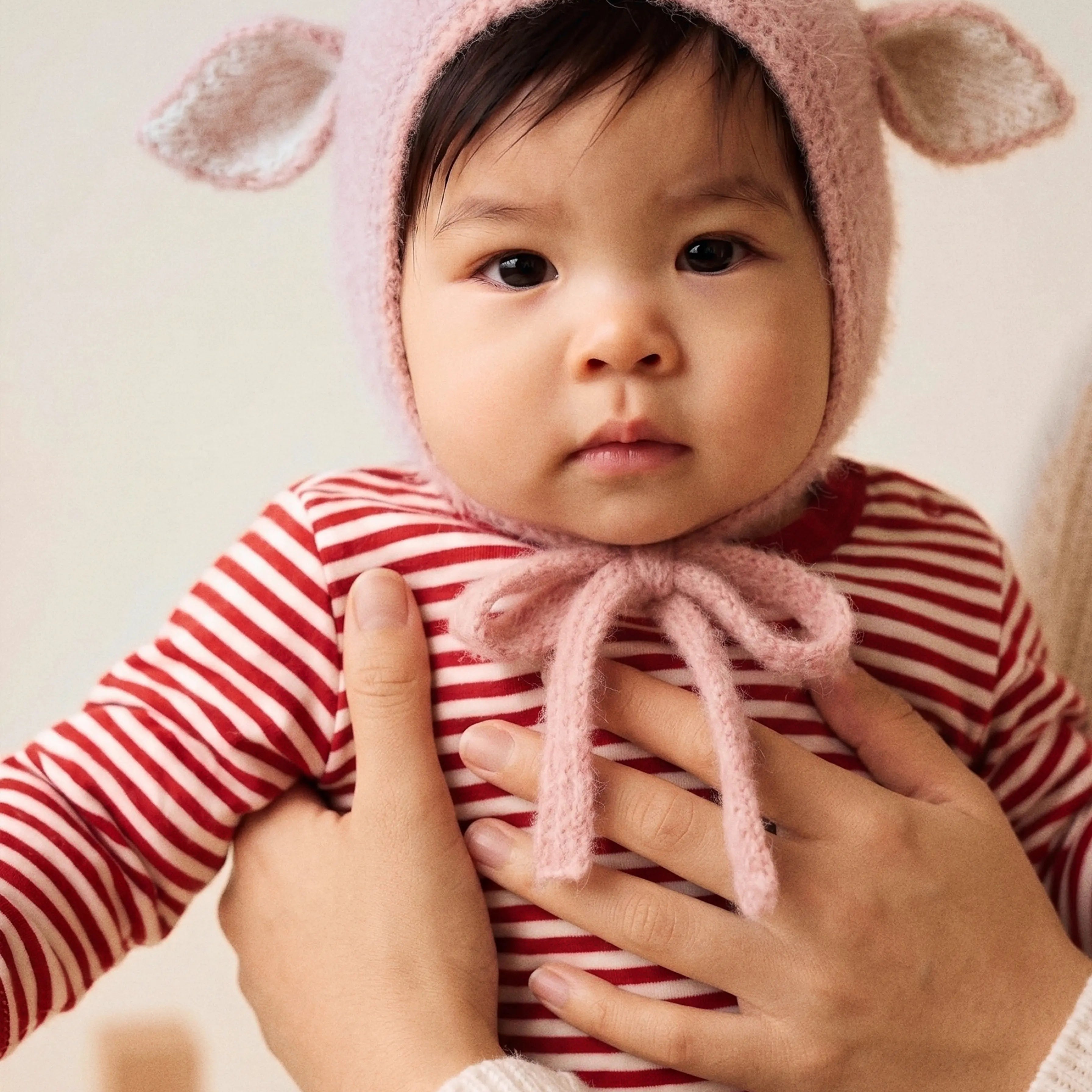 A baby held in a parent's arms wearing the Fairy Hat in Baby Pink, with the soft knitted ties loosely tied under the chin.