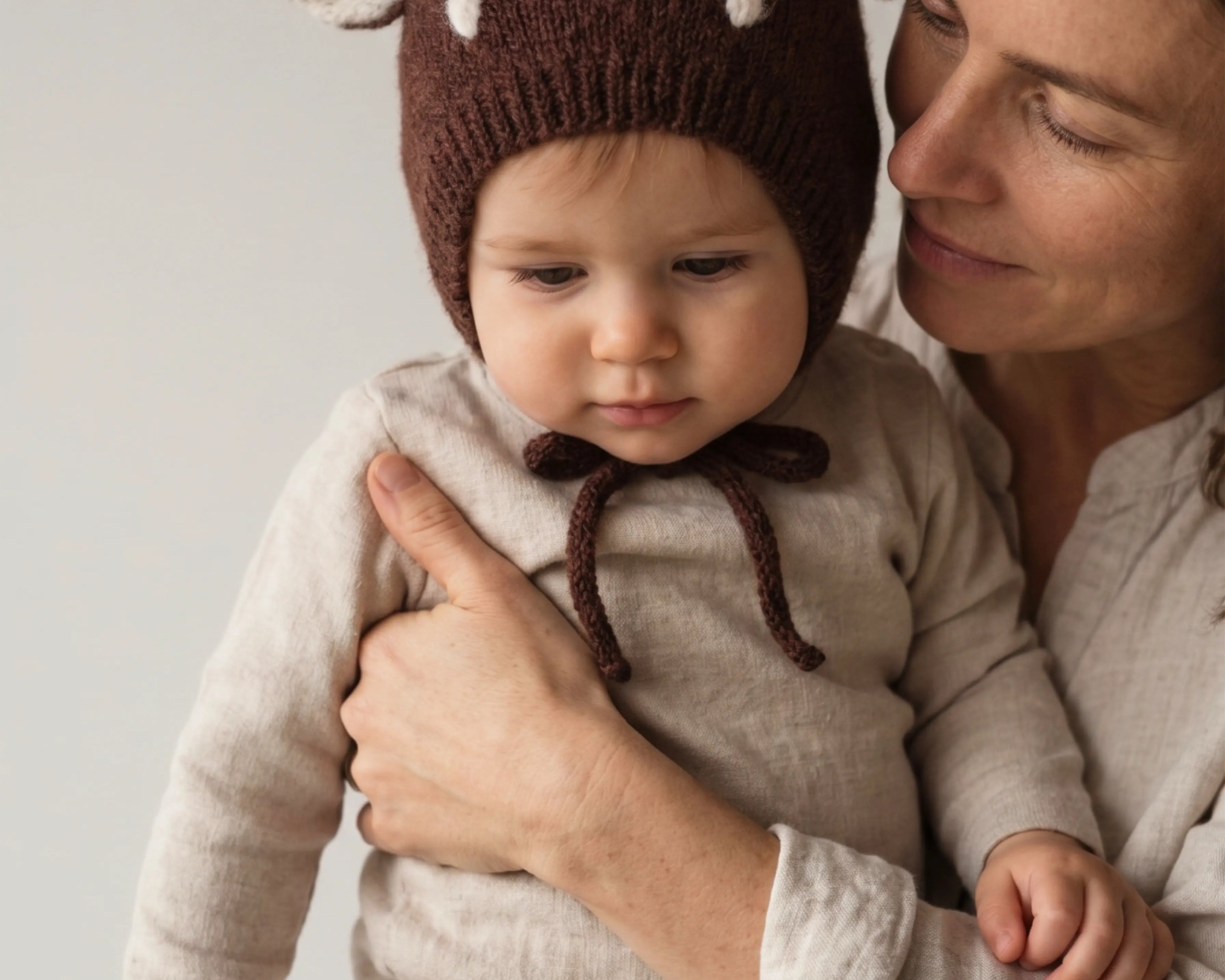 Baby in a knitted bonnet with deer ears and white spots, held close by their mother. 