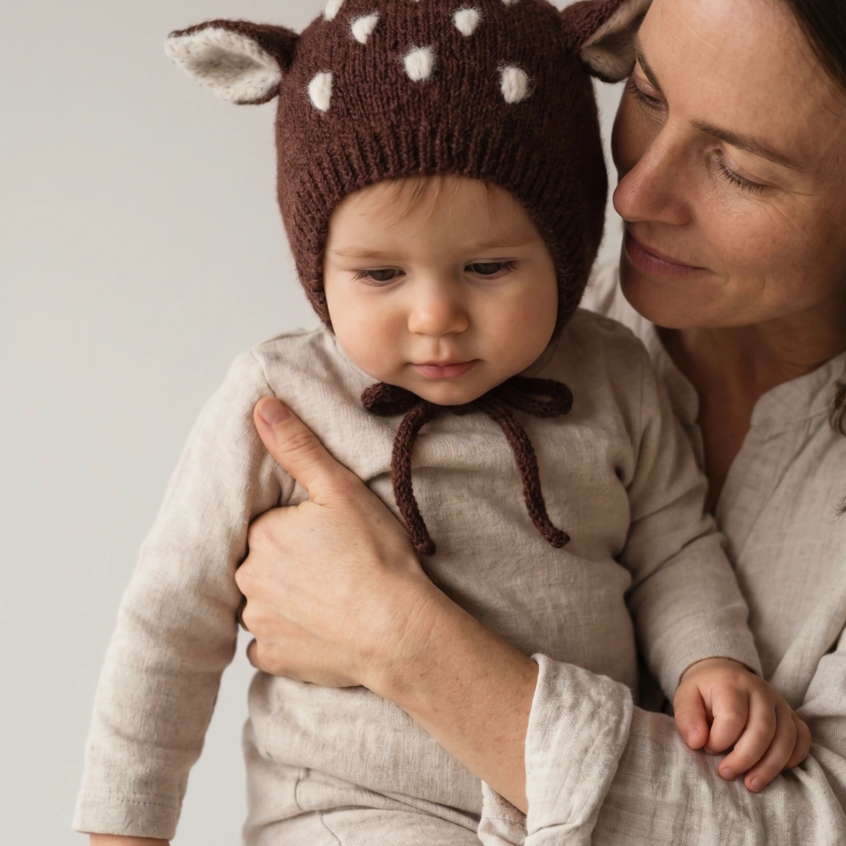Baby in a knitted bonnet with deer ears and white spots, held close by their mother. 