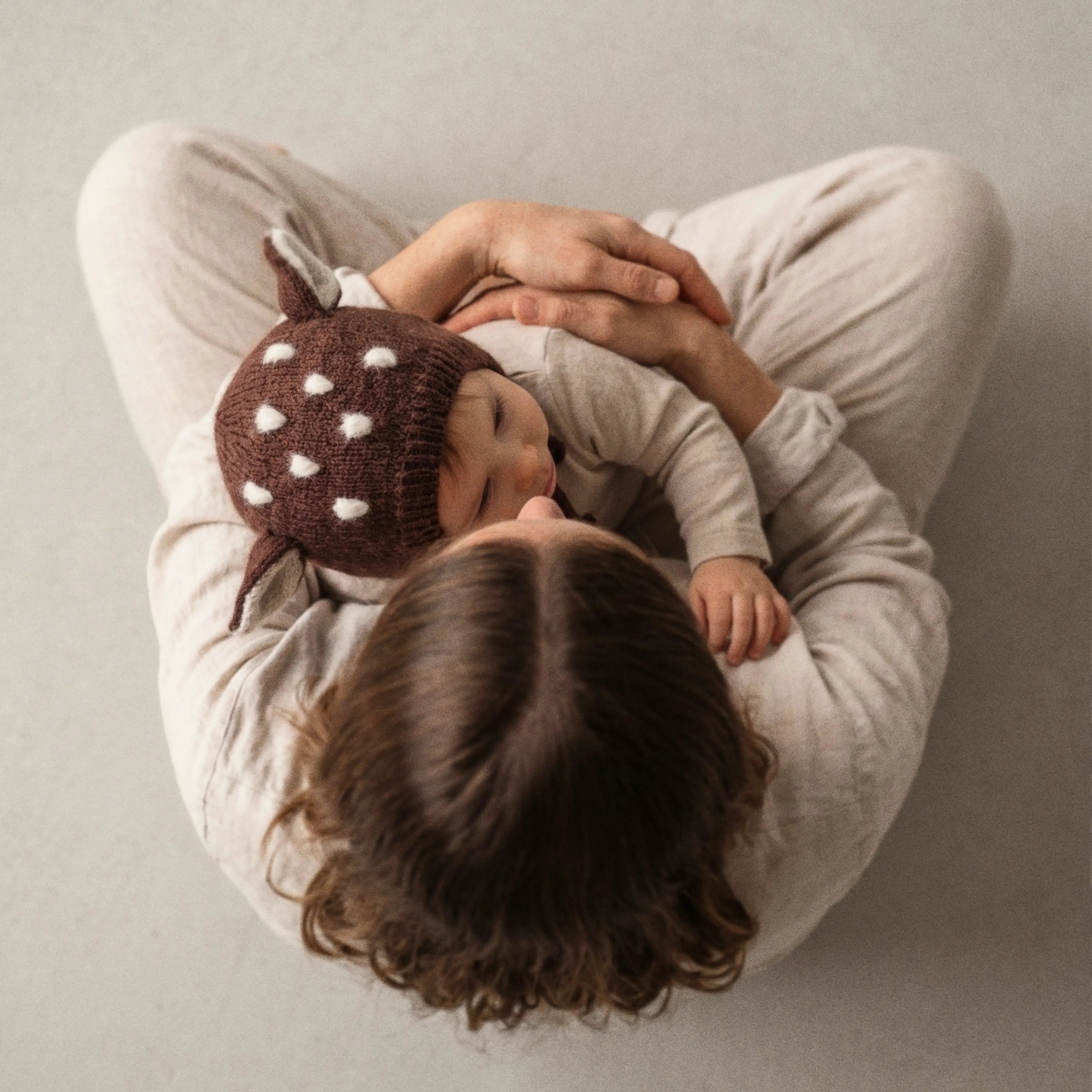 Overhead view of a mother sitting cross-legged holding a baby in a knitted bonnet with deer ears.