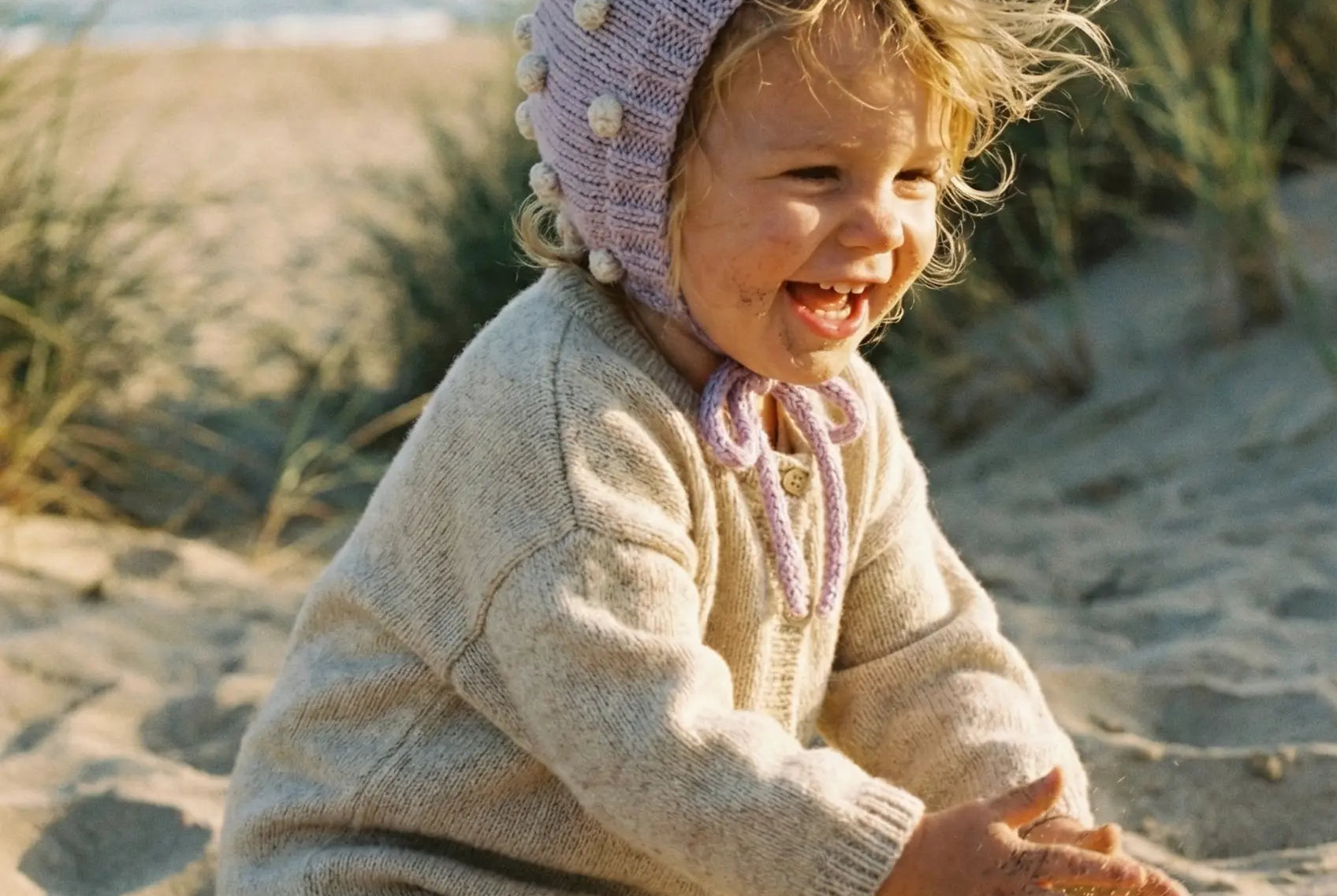 Baby wearing a lavender organic cotton bonnet playing with sand on a beach