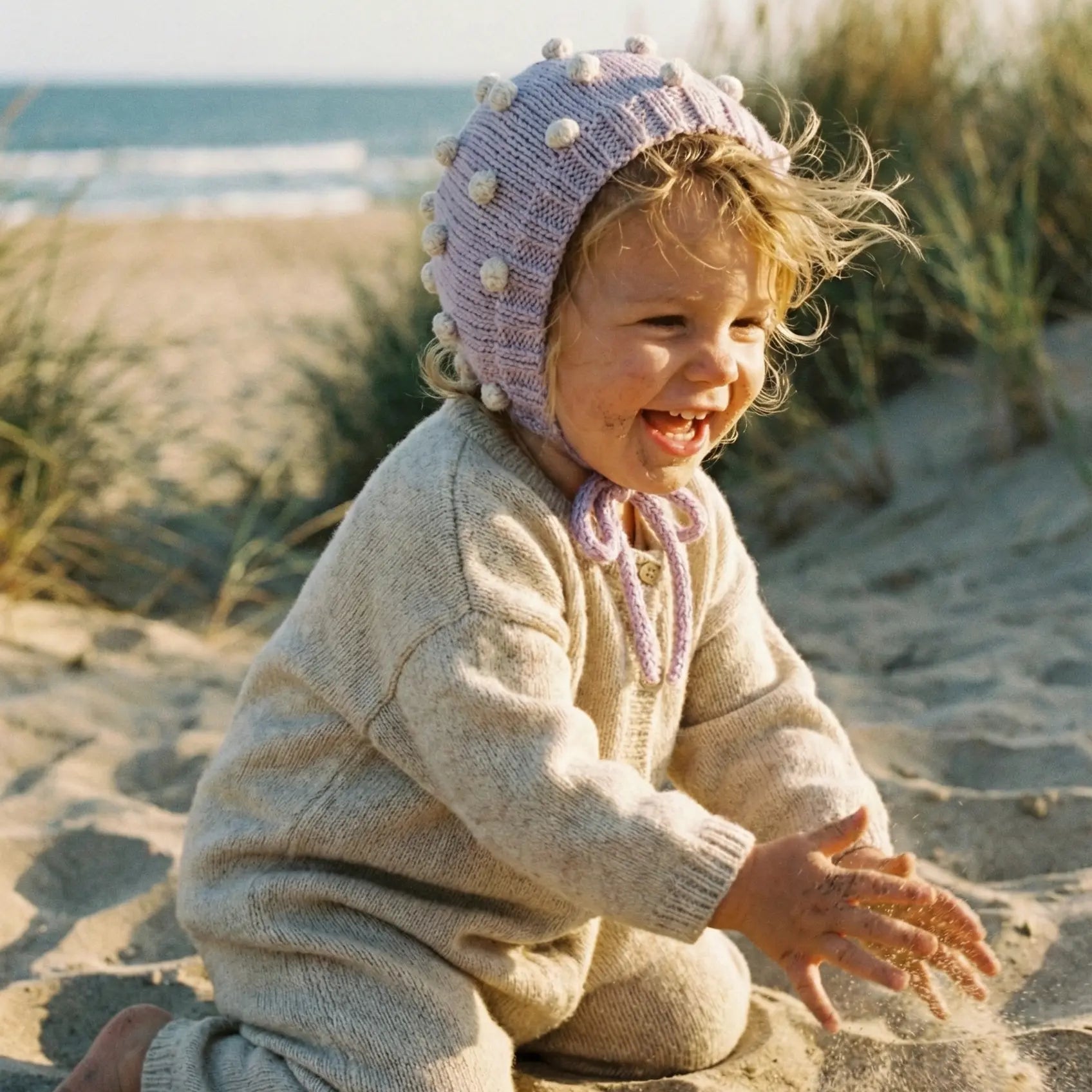 Baby wearing a lavender organic cotton bonnet playing with sand on a beach